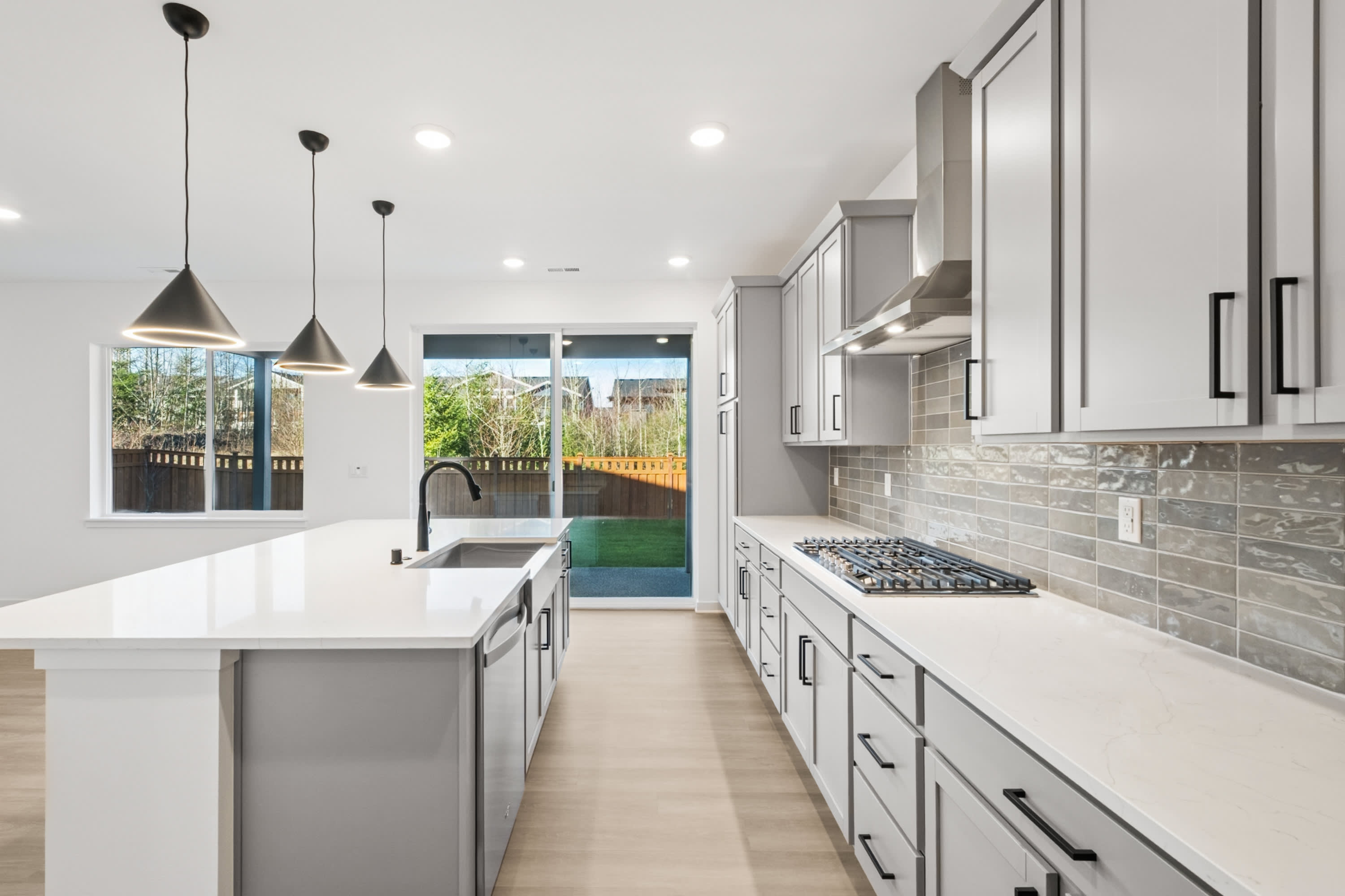 A modern, well-lit kitchen with white cabinets, a stone backsplash, and pendant lights, leading to a backyard visible through a glass door.