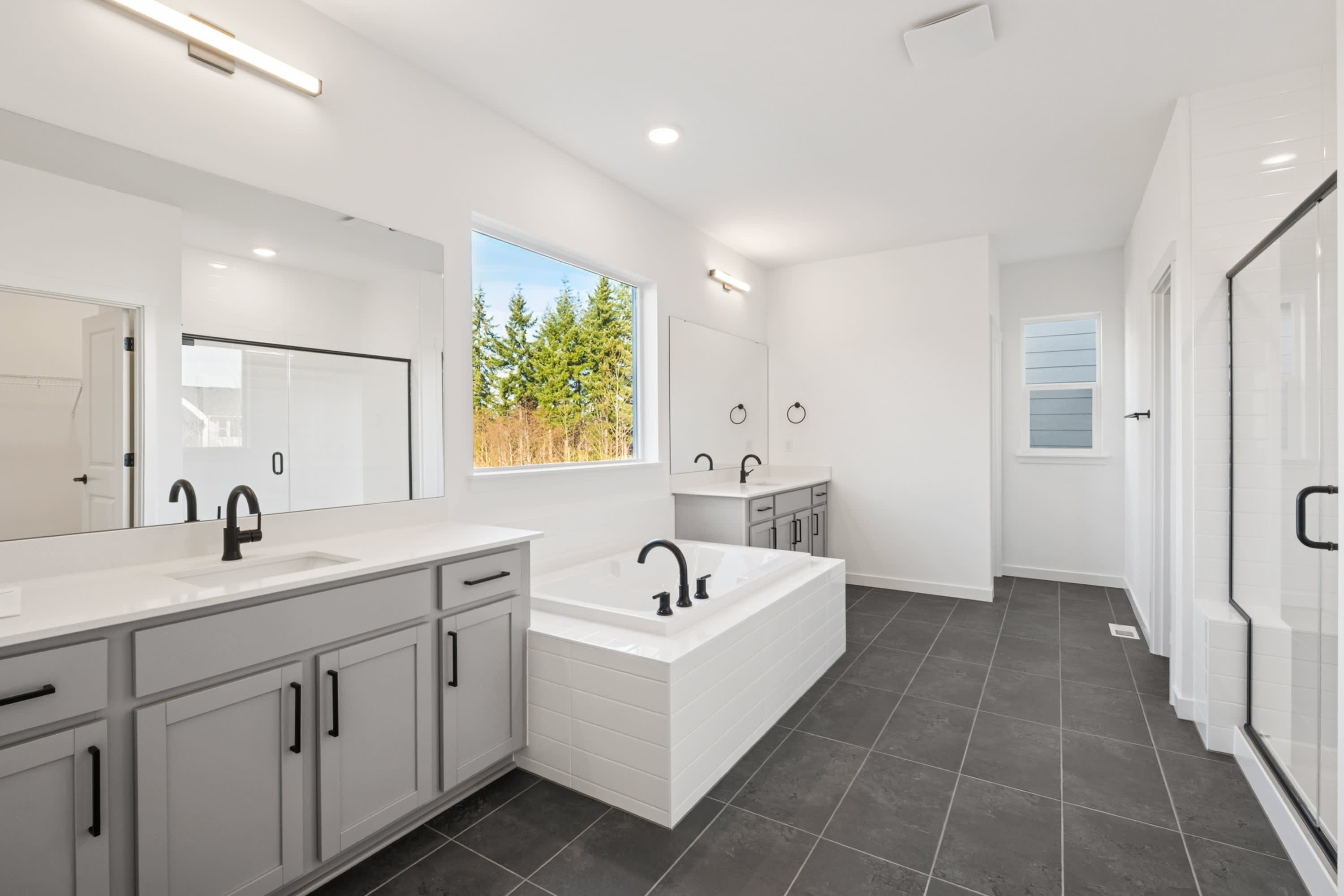 A modern, well-lit bathroom with gray cabinets, a white vanity, and a large window overlooking a wooded area.