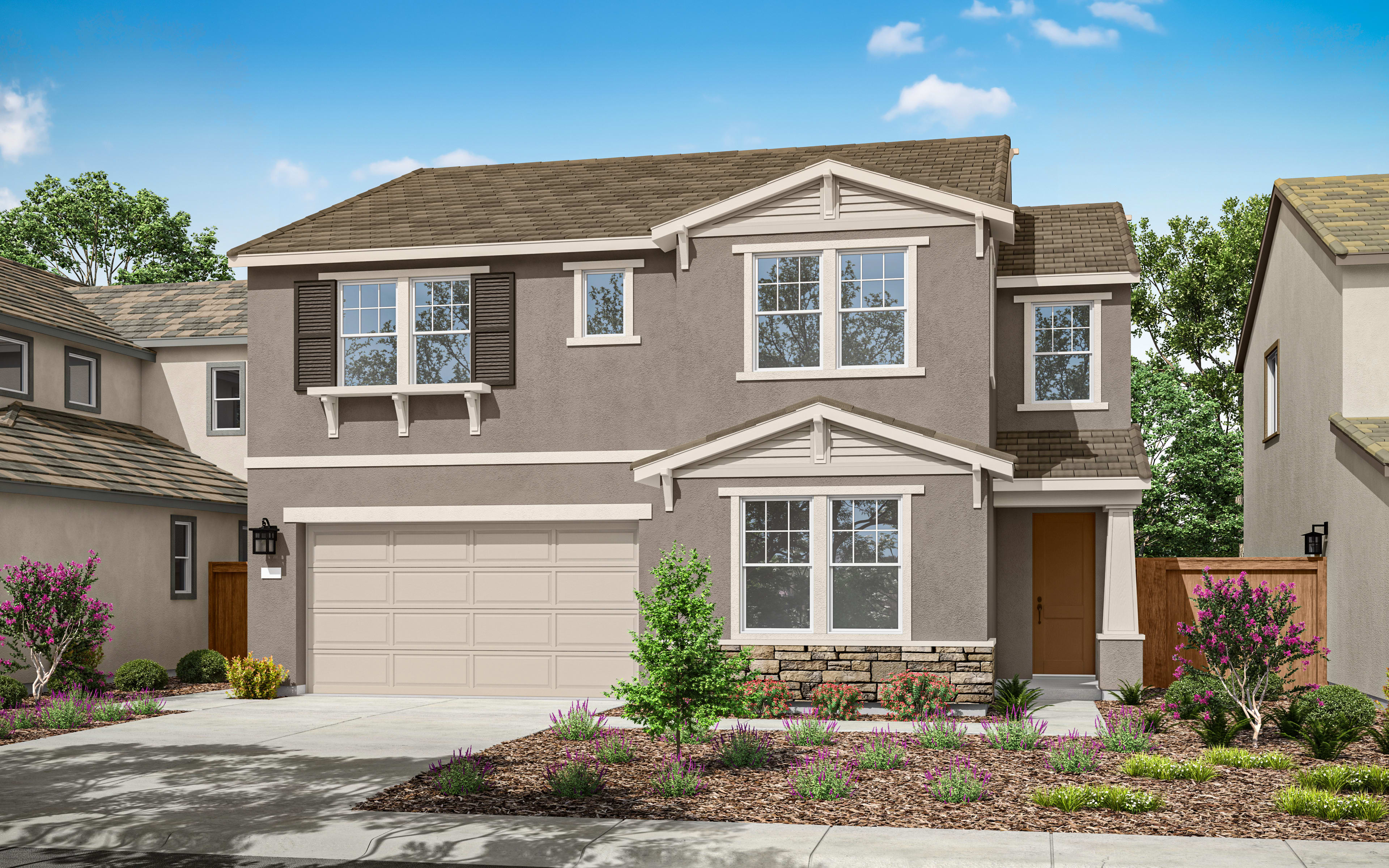 A two-story residential house with a gabled roof, stone accents, and a well-landscaped front yard featuring flowering plants and a paved driveway, set against a backdrop of a clear blue sky with scattered clouds.
