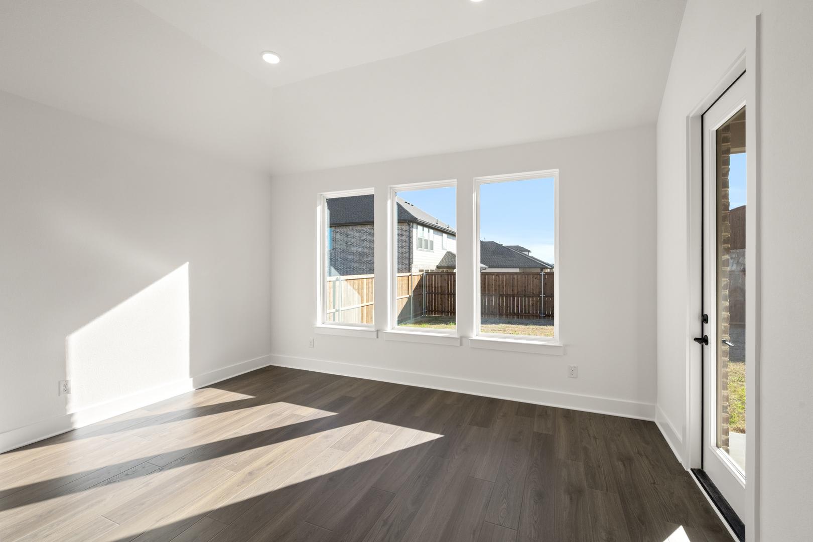 A bright and spacious room with large windows overlooking a backyard, featuring a hardwood floor with a diagonal pattern.