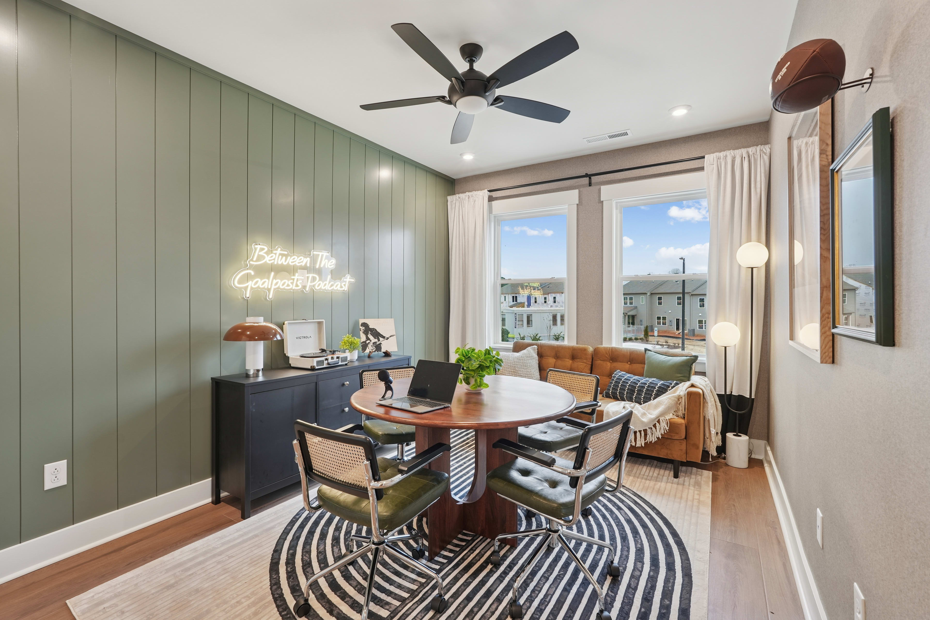 A cozy and well-furnished living room with a dining area, featuring a ceiling fan, large windows, and a striped rug on the hardwood floor.