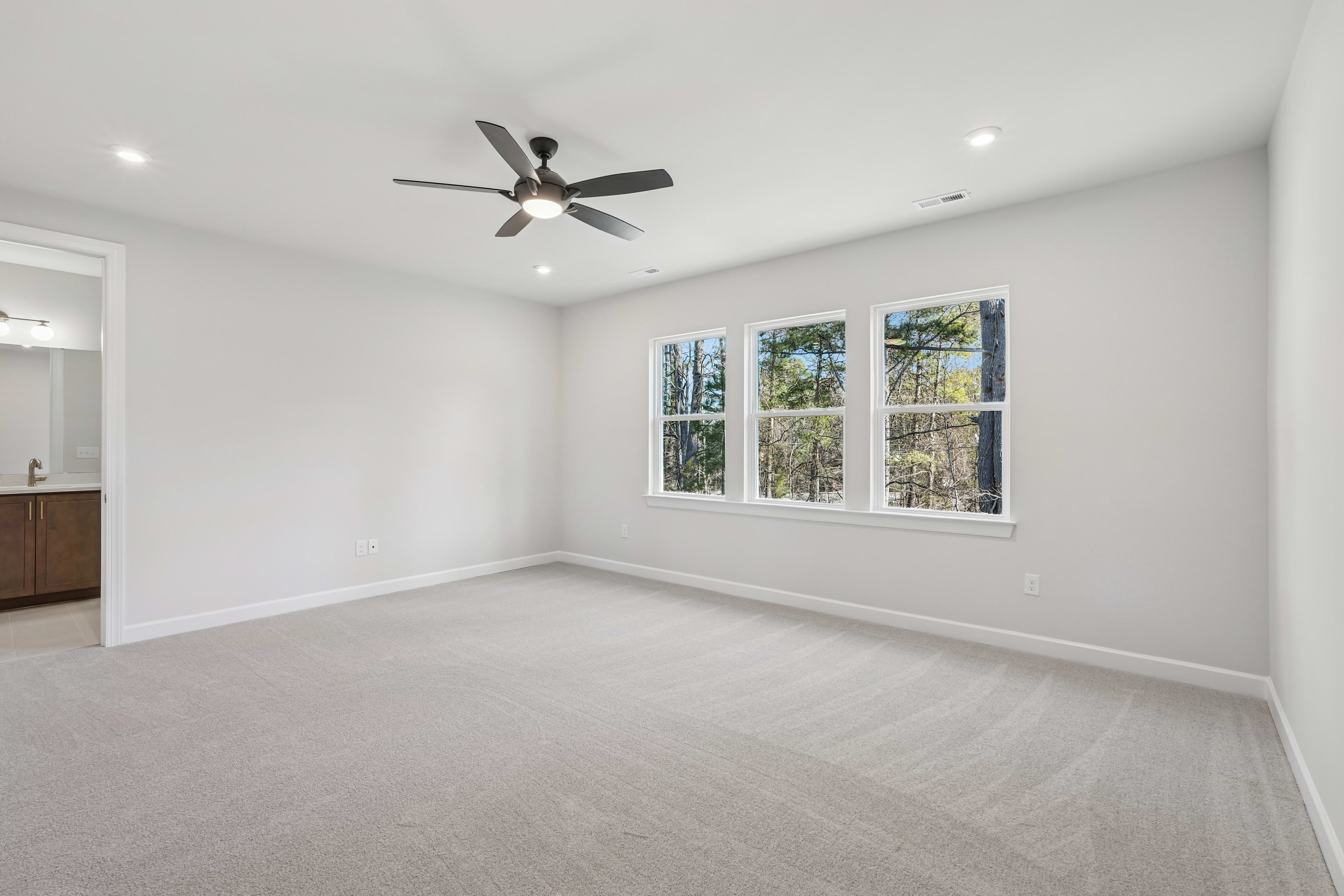A spacious, well-lit room with a ceiling fan, large windows overlooking a wooded area, and a clean, carpeted floor.