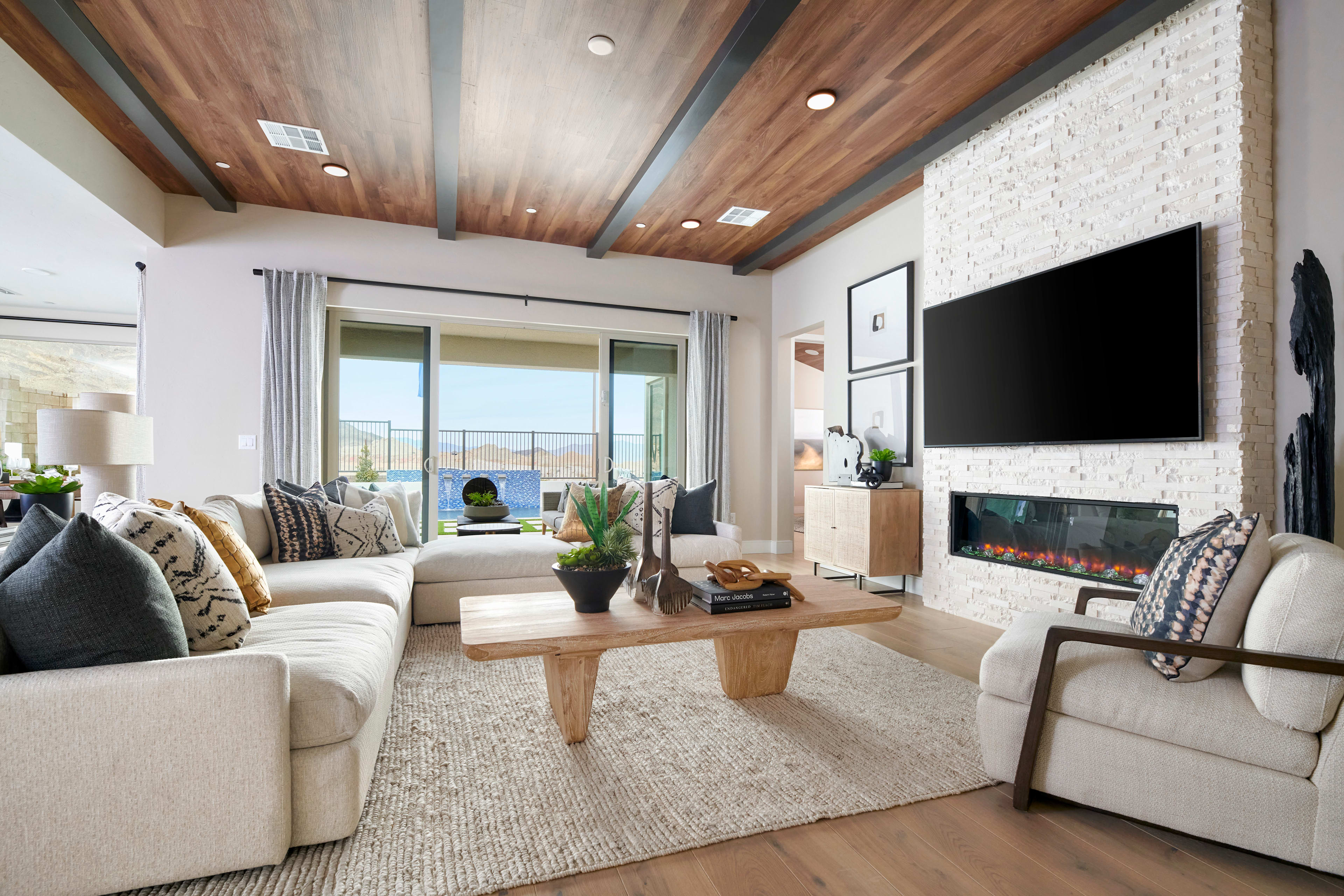A cozy and inviting living room with wooden ceiling beams, a fireplace, and large windows overlooking a scenic outdoor view.