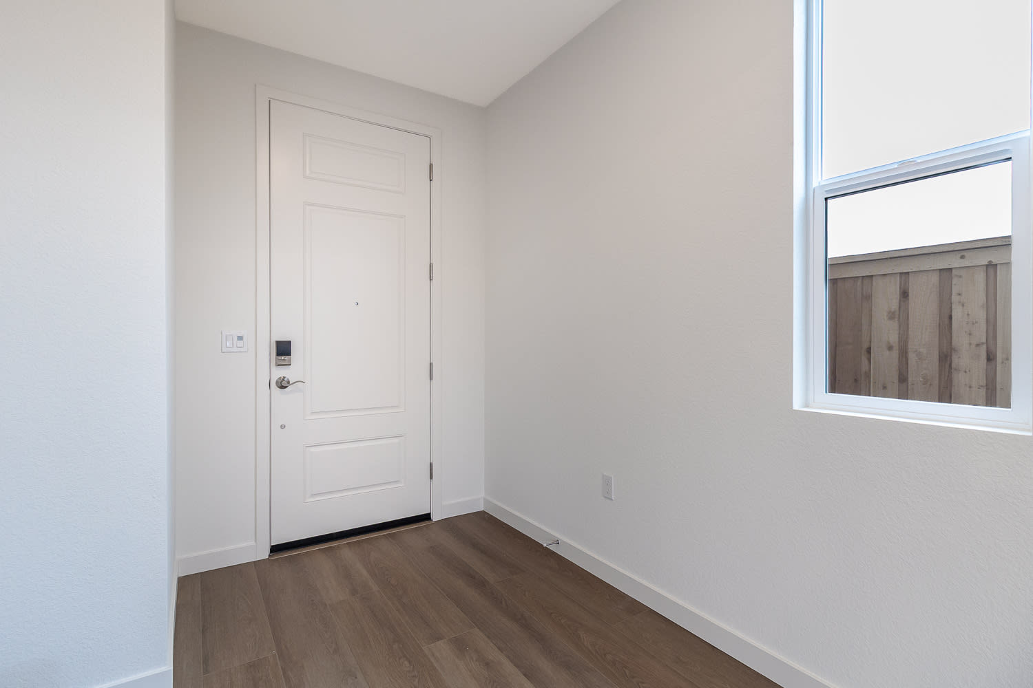 A simple, minimalist interior with a white door, wooden flooring, and a window providing natural light.