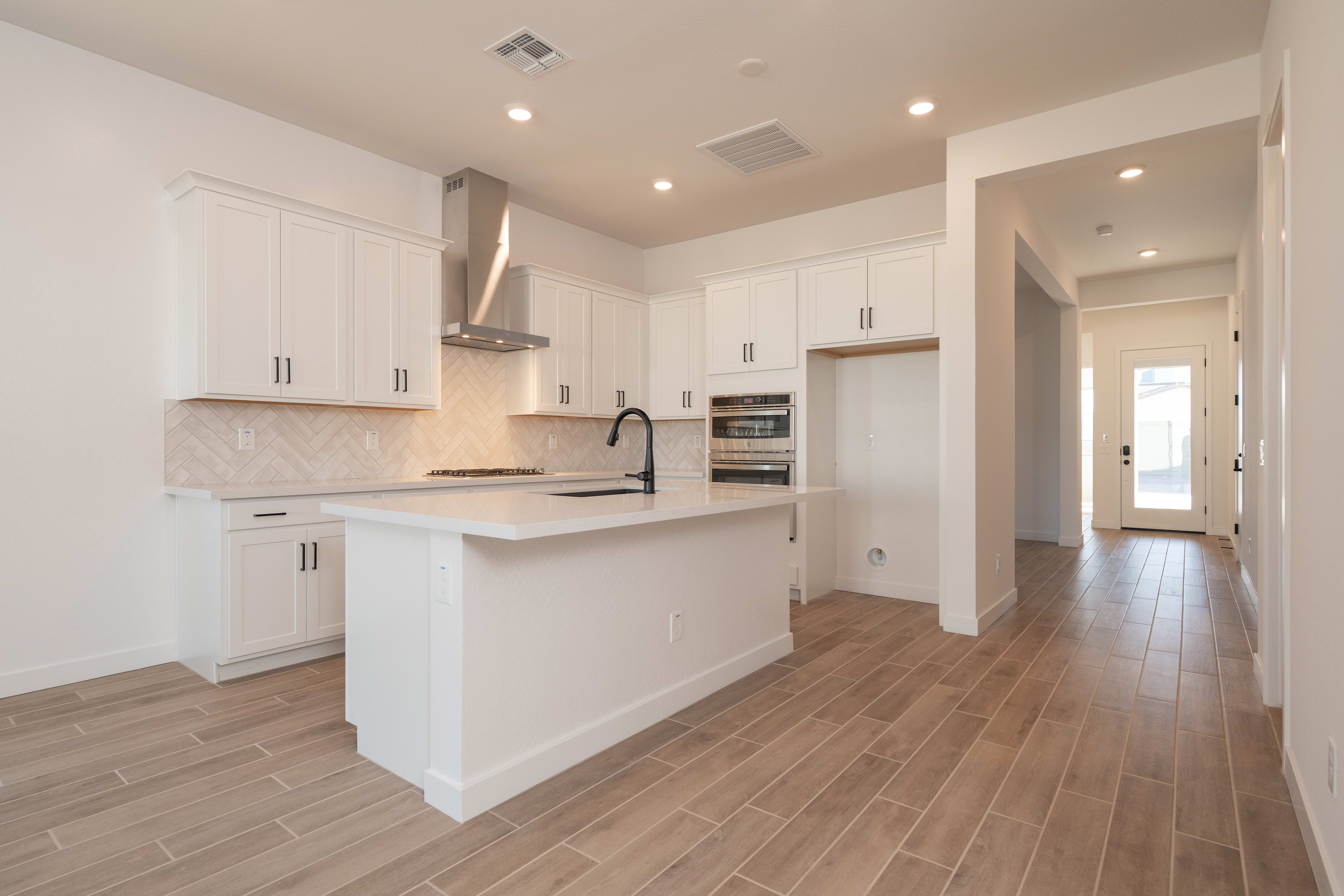 A modern, open-concept kitchen with white cabinets, a tile backsplash, and hardwood flooring extends into a hallway with a wooden floor.