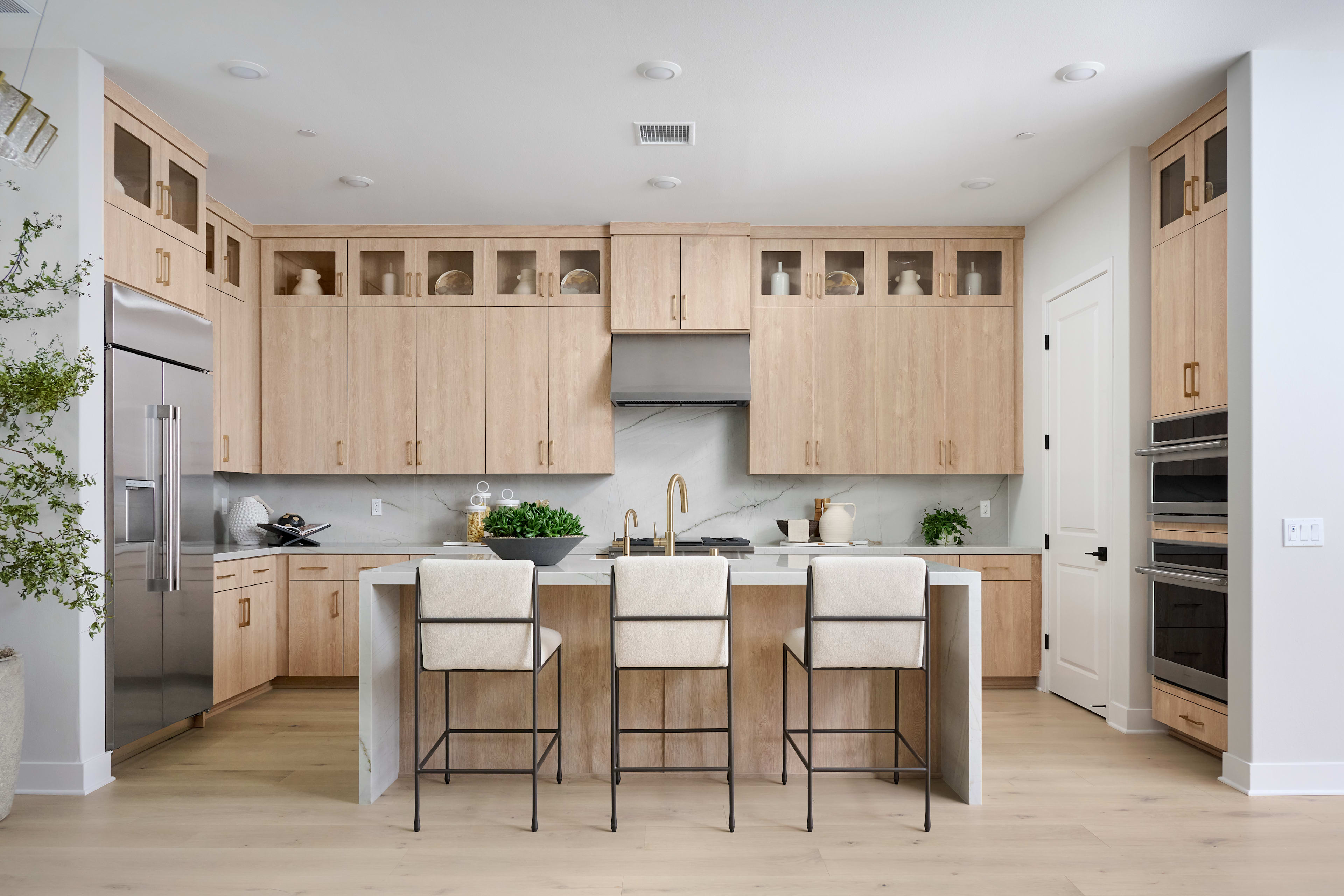 A modern, open-concept kitchen with light-colored wood cabinetry, a central island with bar stools, and stainless steel appliances set against a bright, airy backdrop.