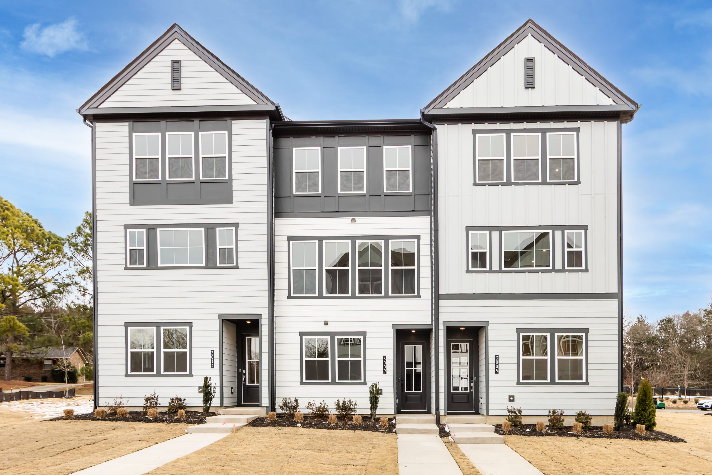 A two-story townhouse with a white exterior, gabled roofs, and large windows stands in a landscaped setting with trees and a clear blue sky in the background.