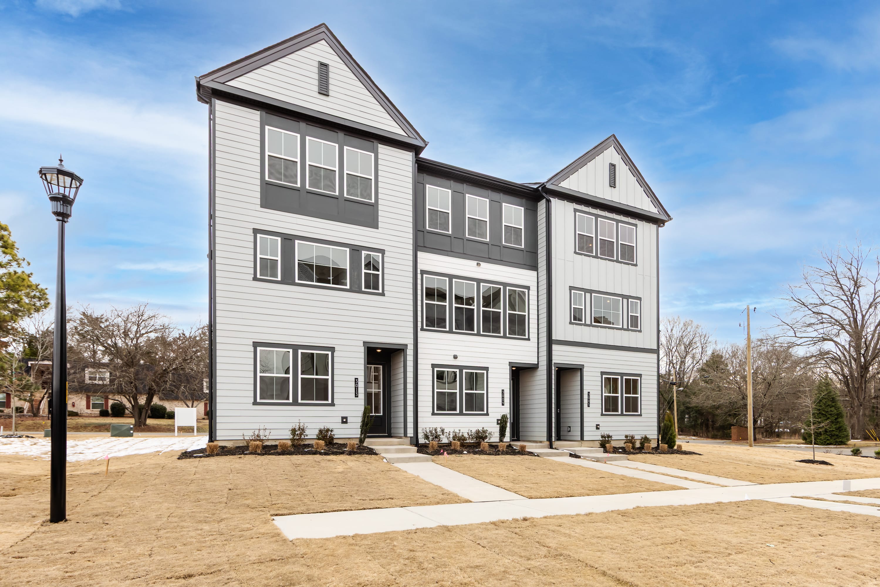A modern, multi-story townhouse with a gray exterior stands in a well-landscaped area, with a paved walkway leading to the entrance and a street lamp visible in the background.