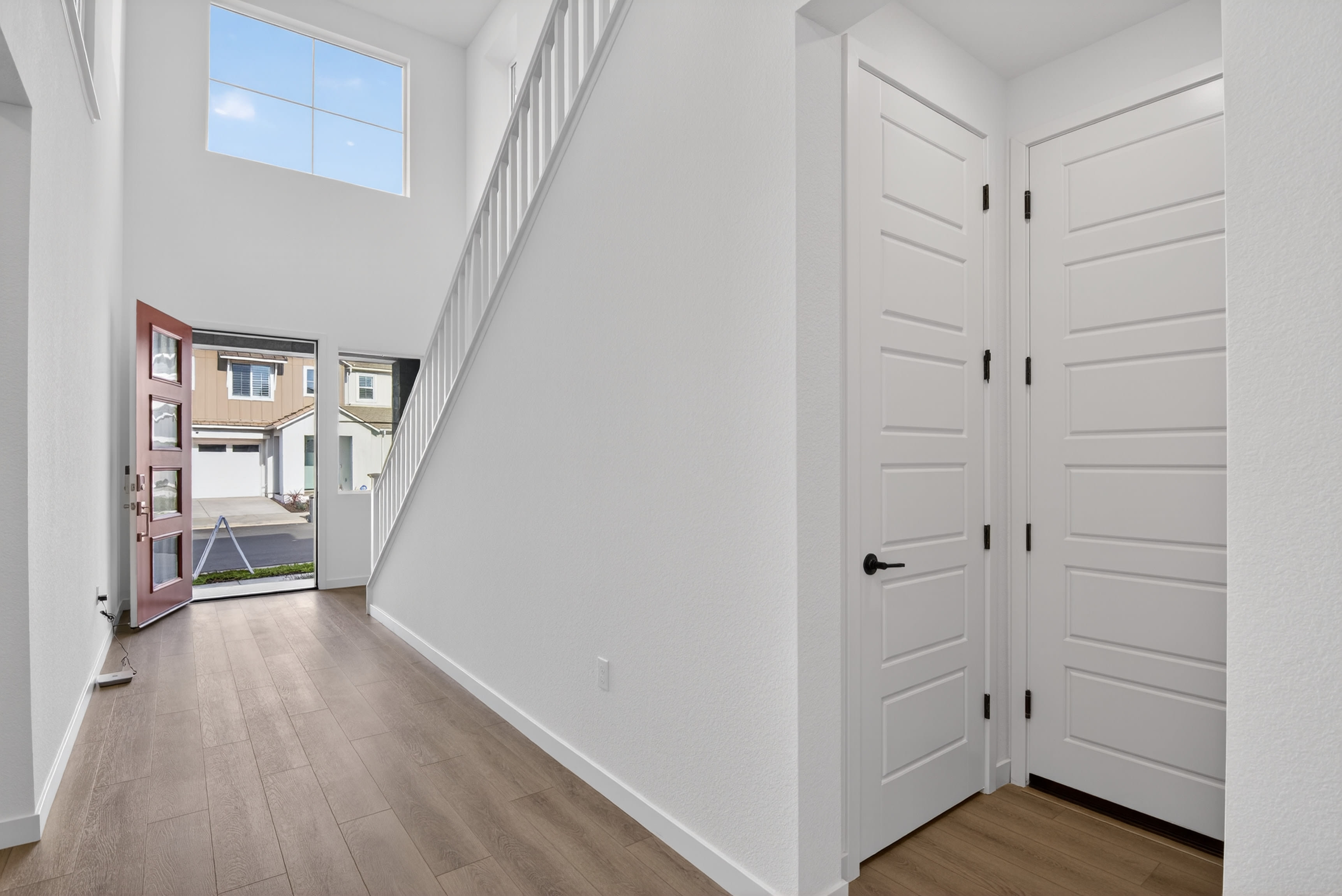 A bright, spacious hallway with white walls, wooden floors, and a window letting in natural light. The hallway leads to a partially visible exterior through an open door.