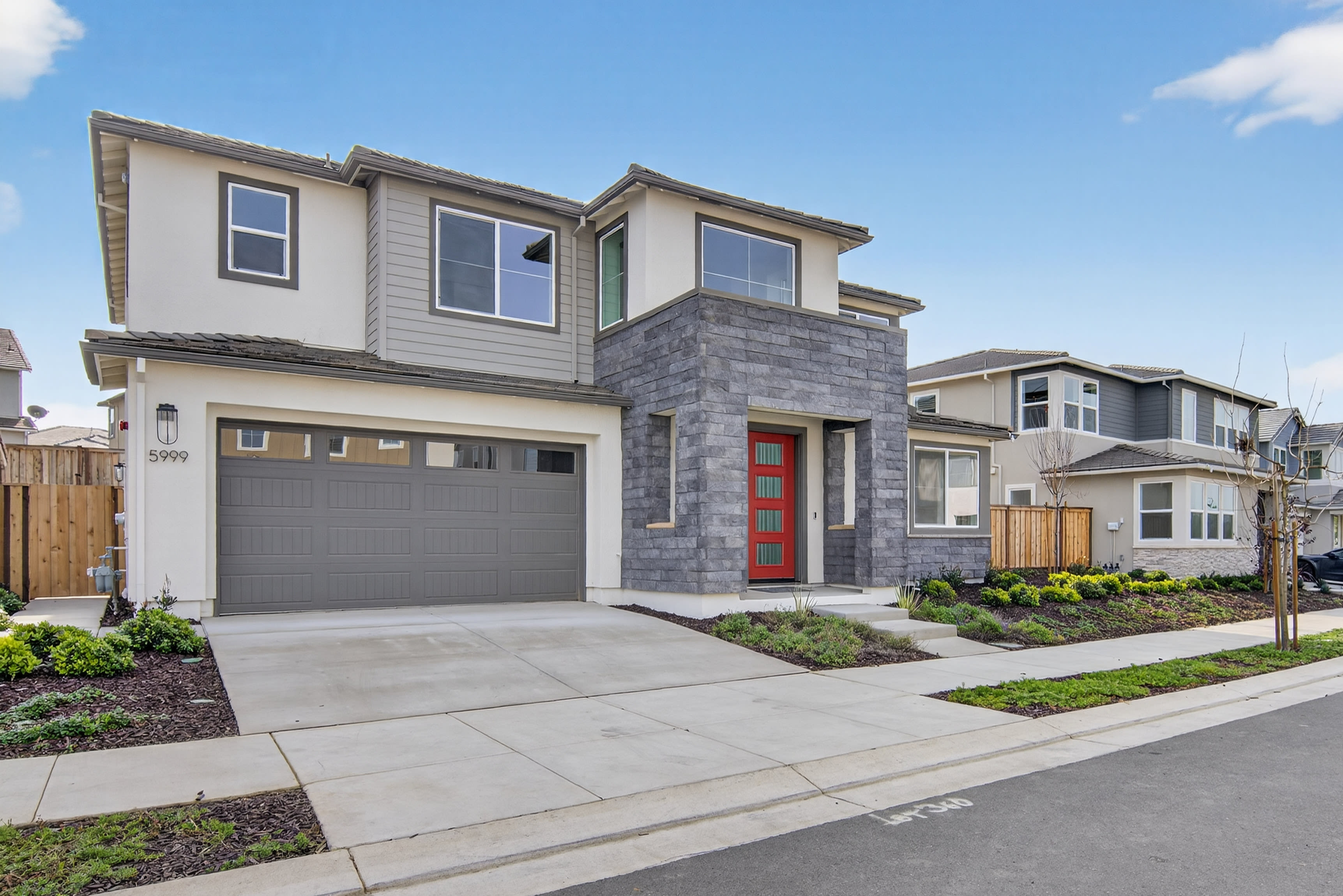 A modern two-story residential house with a gray exterior, a red front door, and a well-landscaped yard in the foreground, set against a clear blue sky with some clouds.