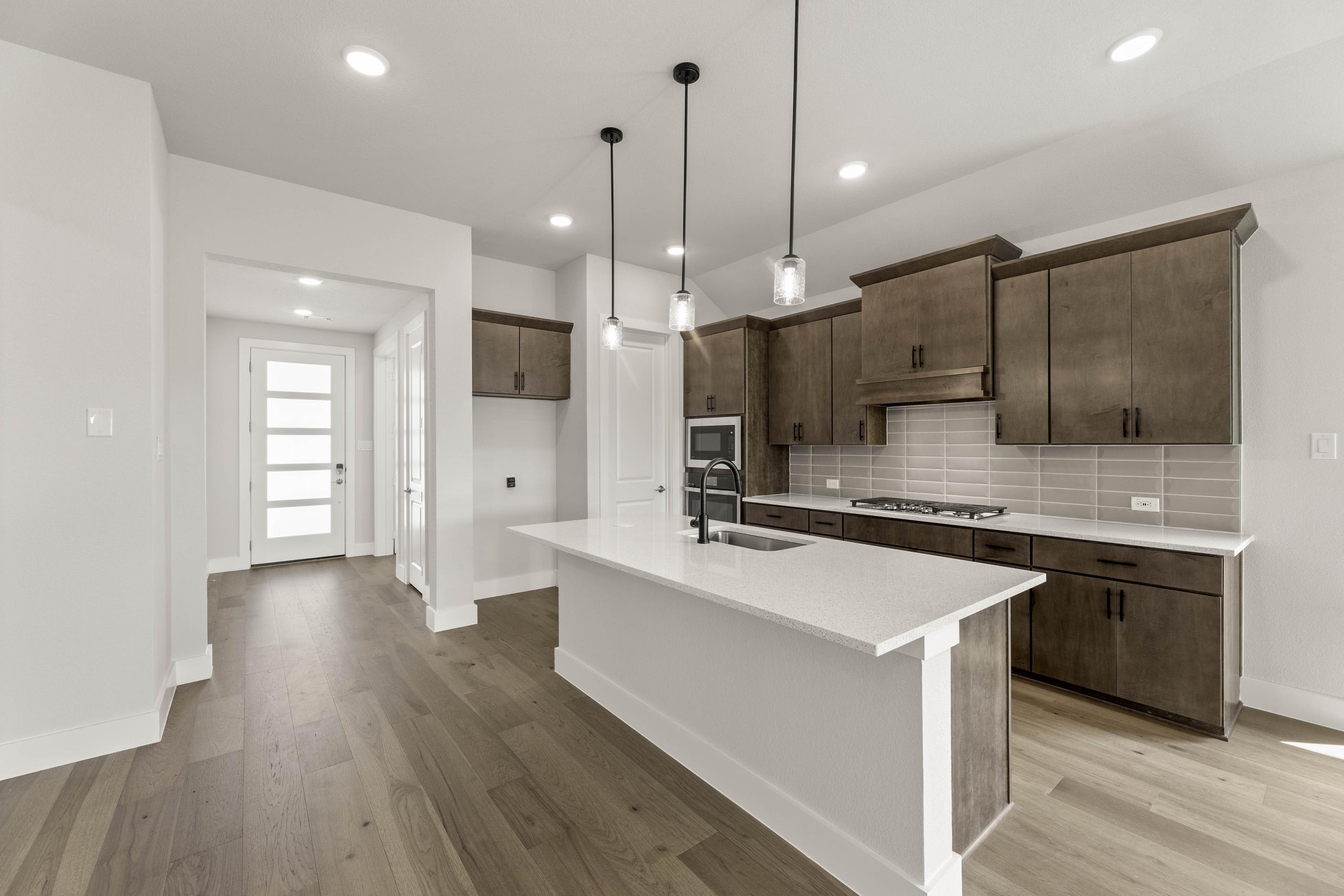 A modern, open-concept kitchen with dark wood cabinets, a white countertop, and pendant lighting fixtures, leading into a hallway with a white door.