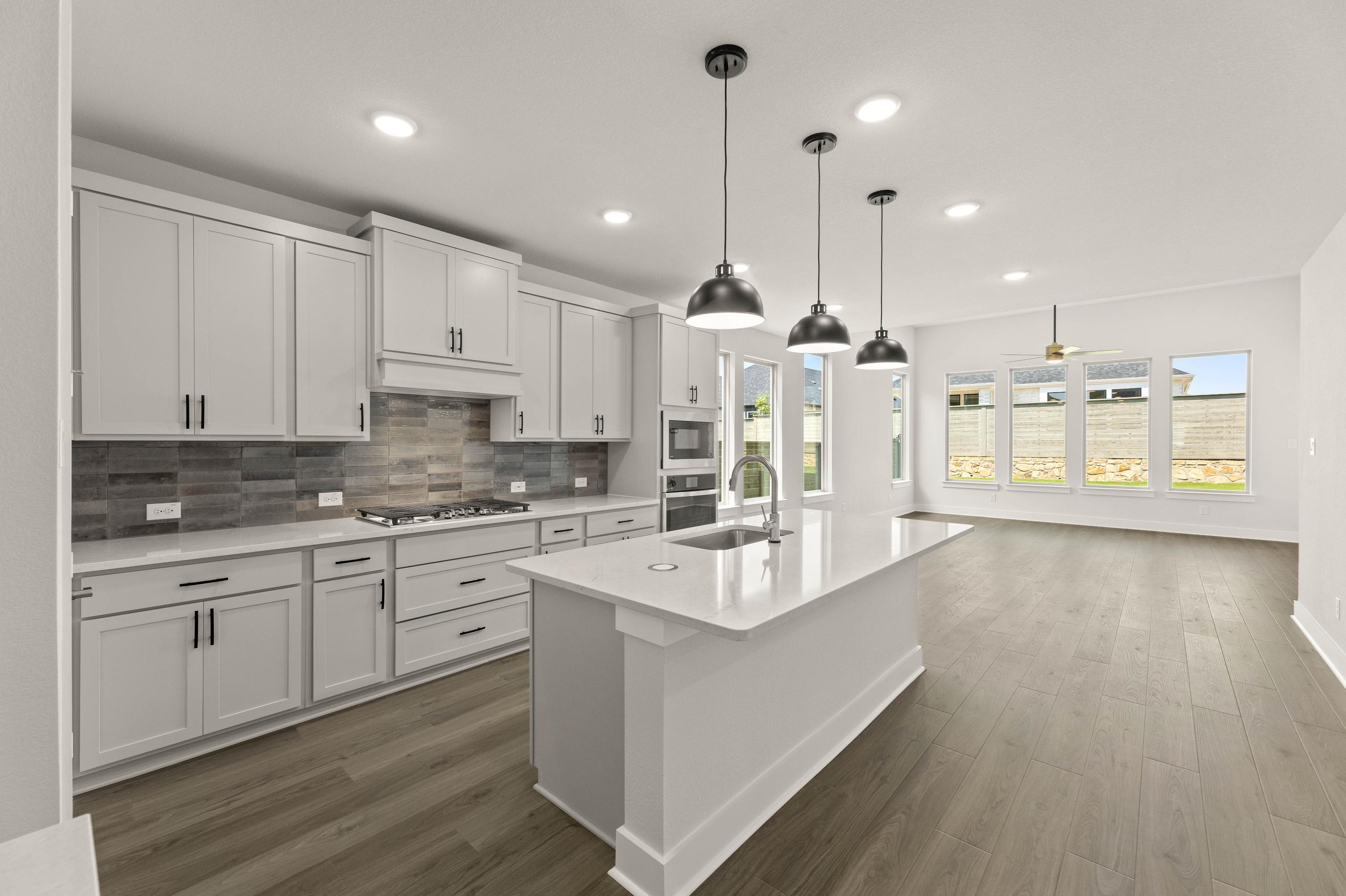 A modern, open-concept kitchen with white cabinets, gray tile backsplash, and pendant lighting, leading into a bright and airy living space with hardwood floors.