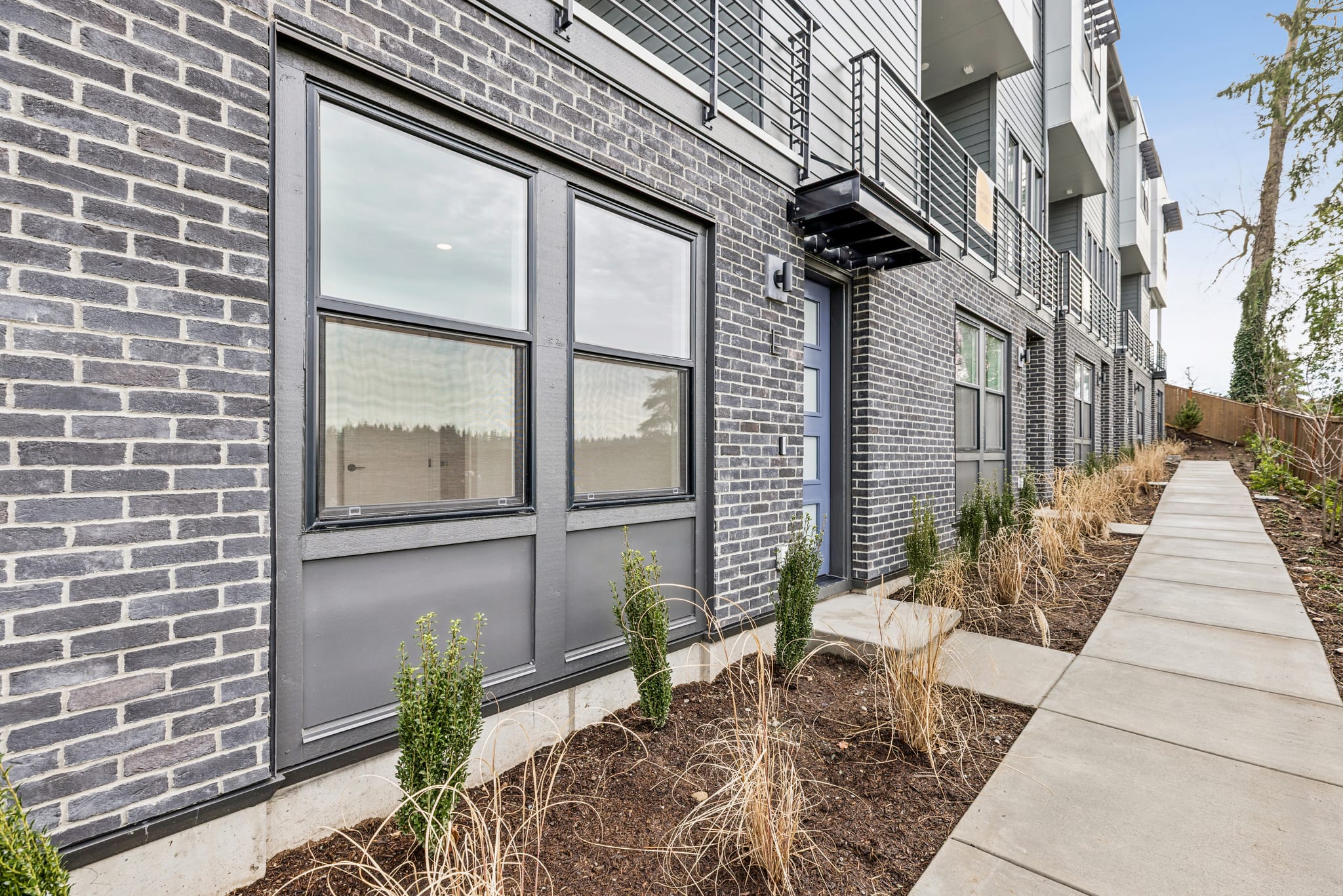 A modern brick building with large windows and a walkway in the foreground, surrounded by landscaping and vegetation.