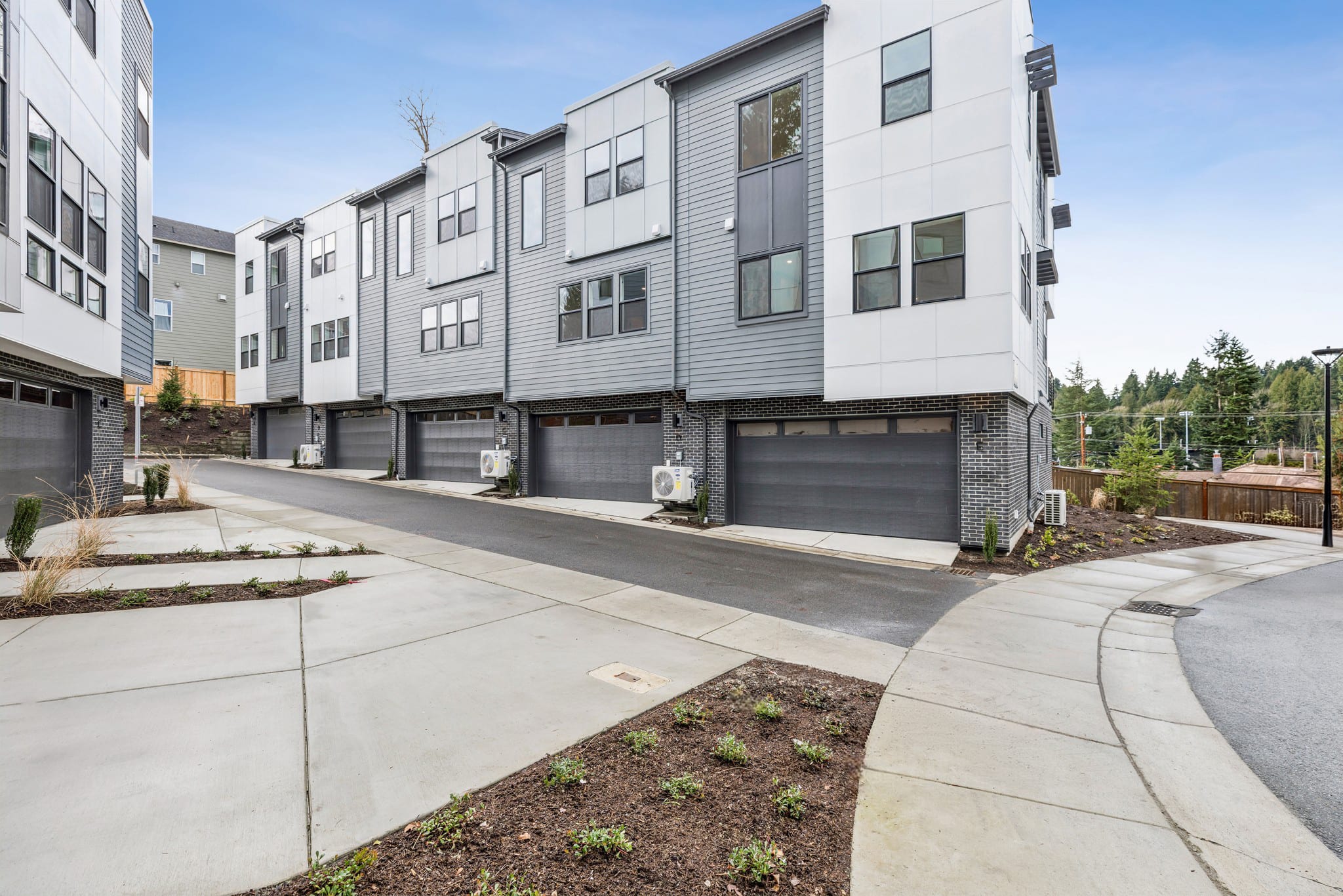 A modern multi-story residential building with attached garages, surrounded by a paved walkway and landscaped areas in the foreground.
