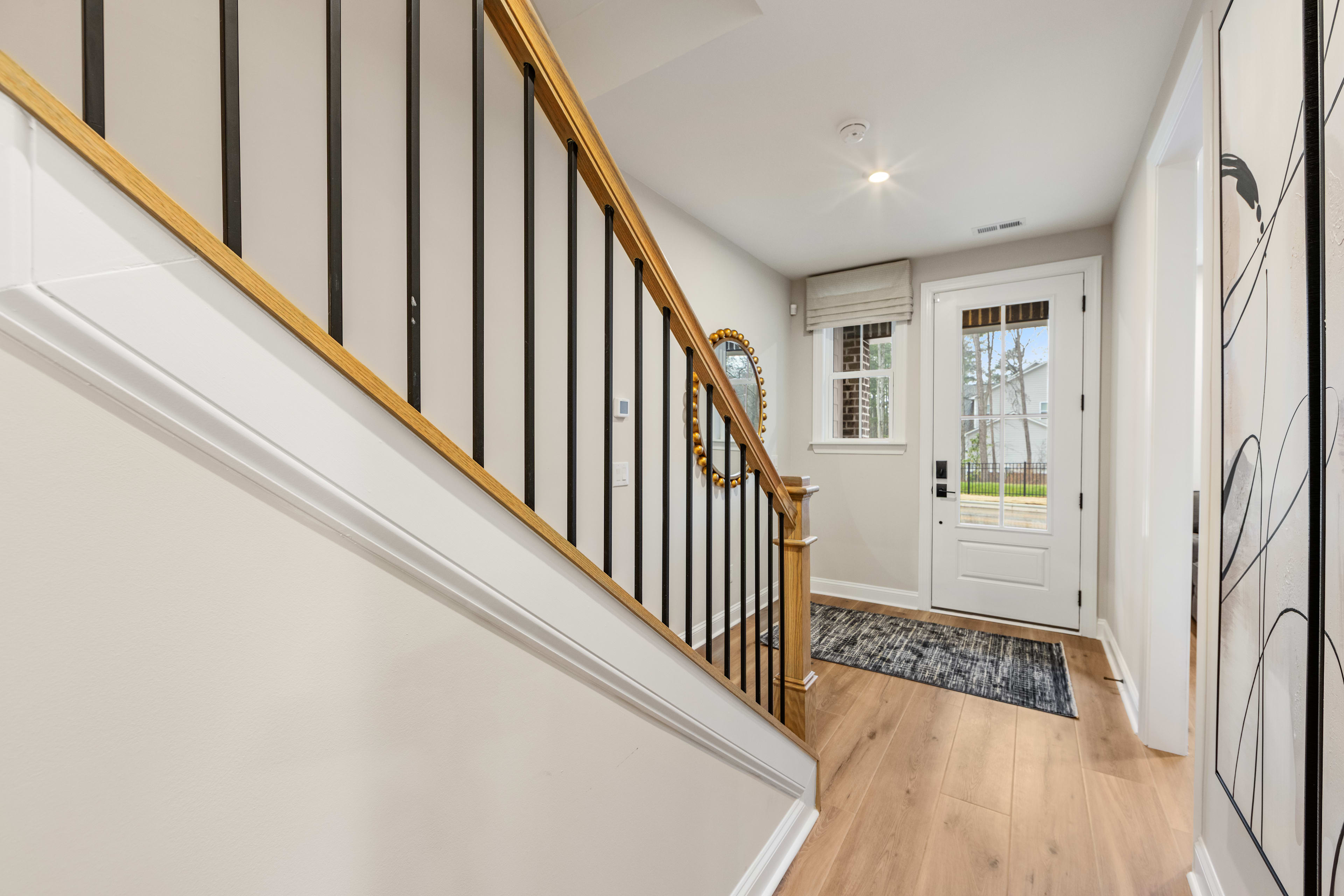 A bright and airy hallway with a wooden staircase, white walls, and a patterned rug leading to a glass-paneled door that provides a view of the outdoors.