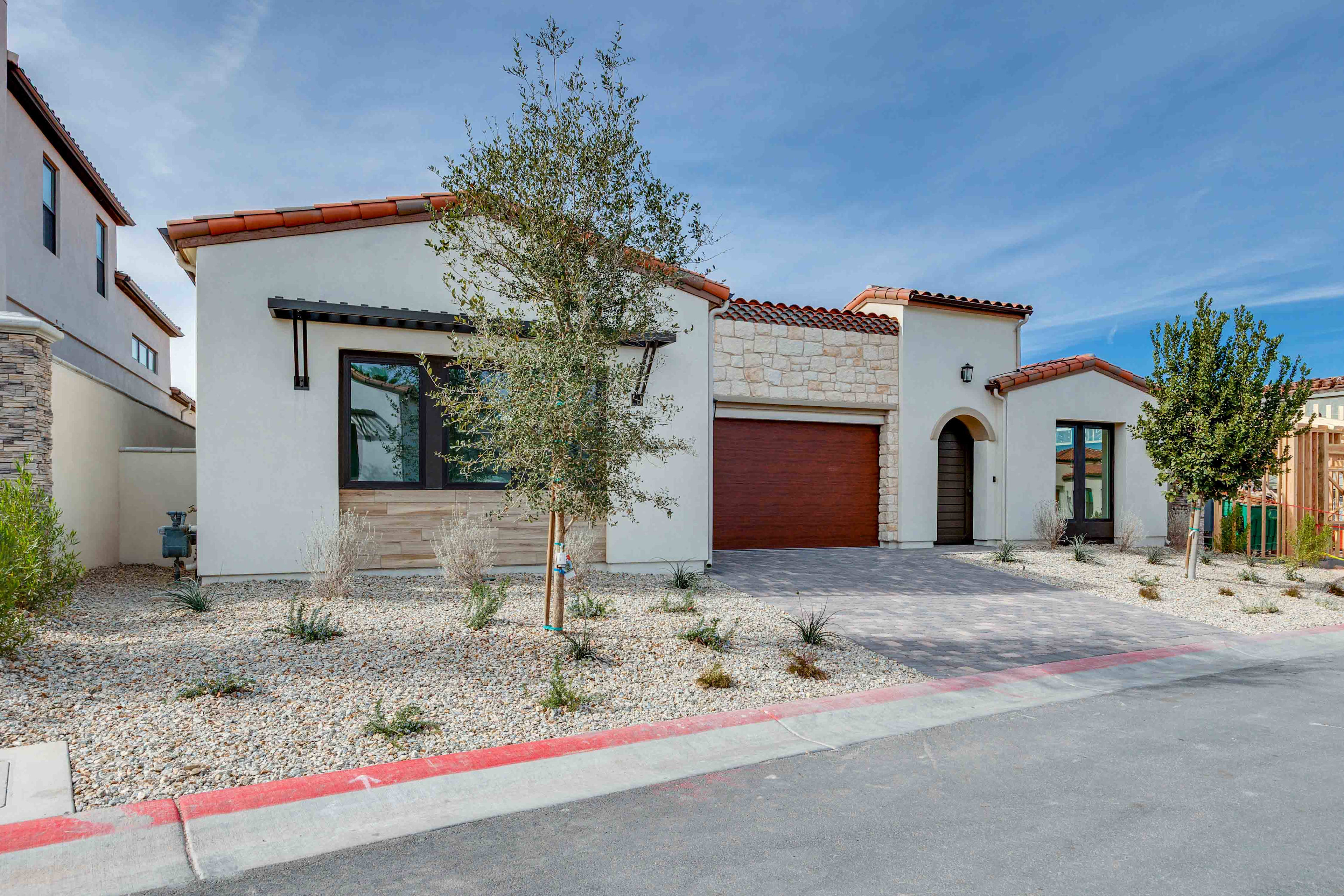 A modern, Mediterranean-style house with a red tile roof, surrounded by a paved driveway and landscaped with a small tree and desert plants in the foreground, set against a clear blue sky.