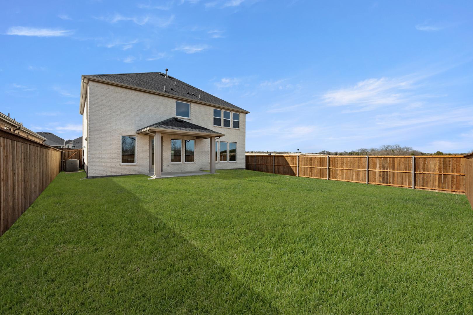A well-manicured lawn surrounds a two-story house with a wooden fence in the background, set against a clear blue sky.