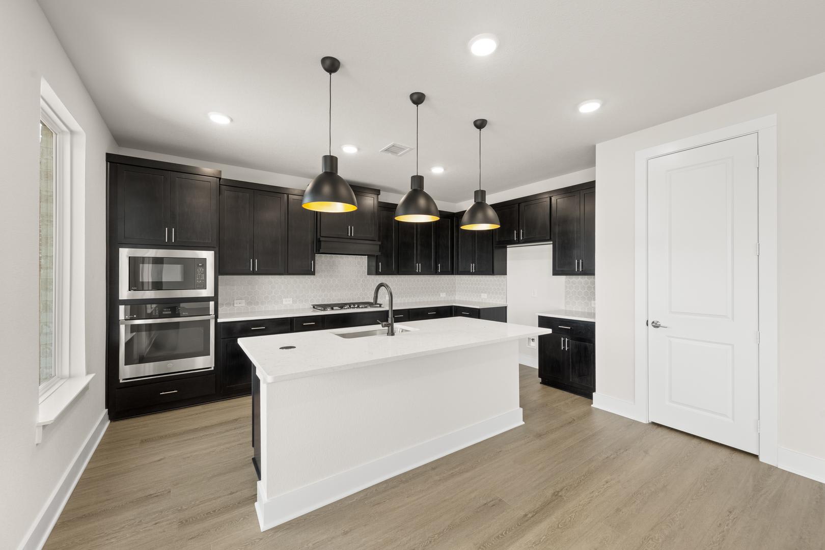 A modern, open-concept kitchen with dark cabinets, a white island, and pendant lights hanging above, set against a light-colored hardwood floor.