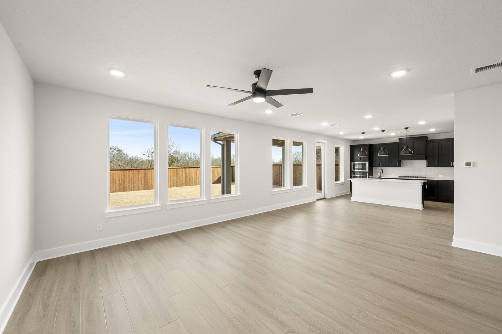 A spacious and modern open-plan living area with a ceiling fan, large windows, and a kitchen visible in the background.