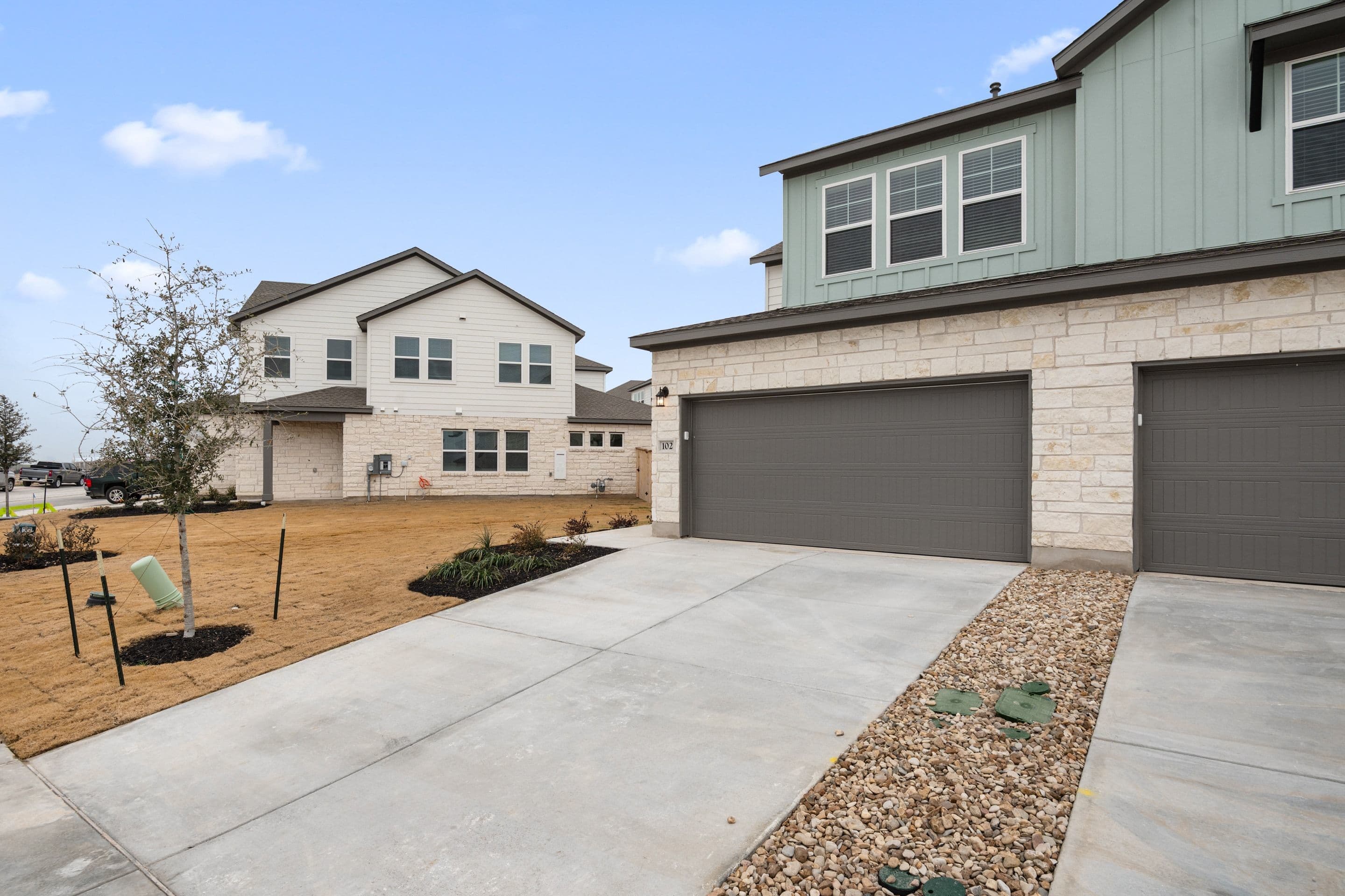 A newly constructed two-story residential building with a garage, surrounded by a paved driveway and landscaped yard with gravel and plants.