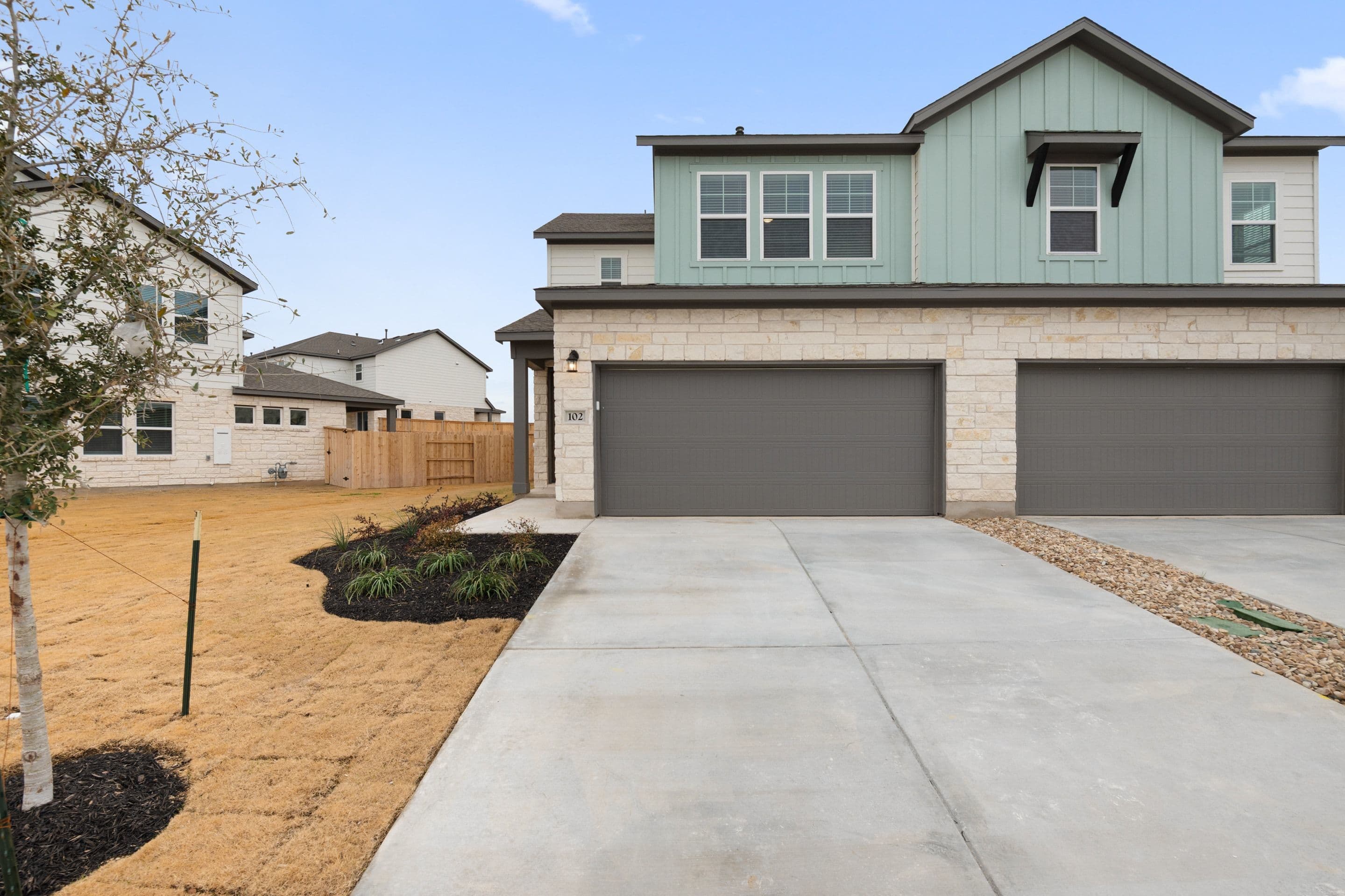 A two-story house with a gray exterior, a garage door, and a concrete driveway leading to the front entrance, surrounded by landscaping and other residential buildings in the background.