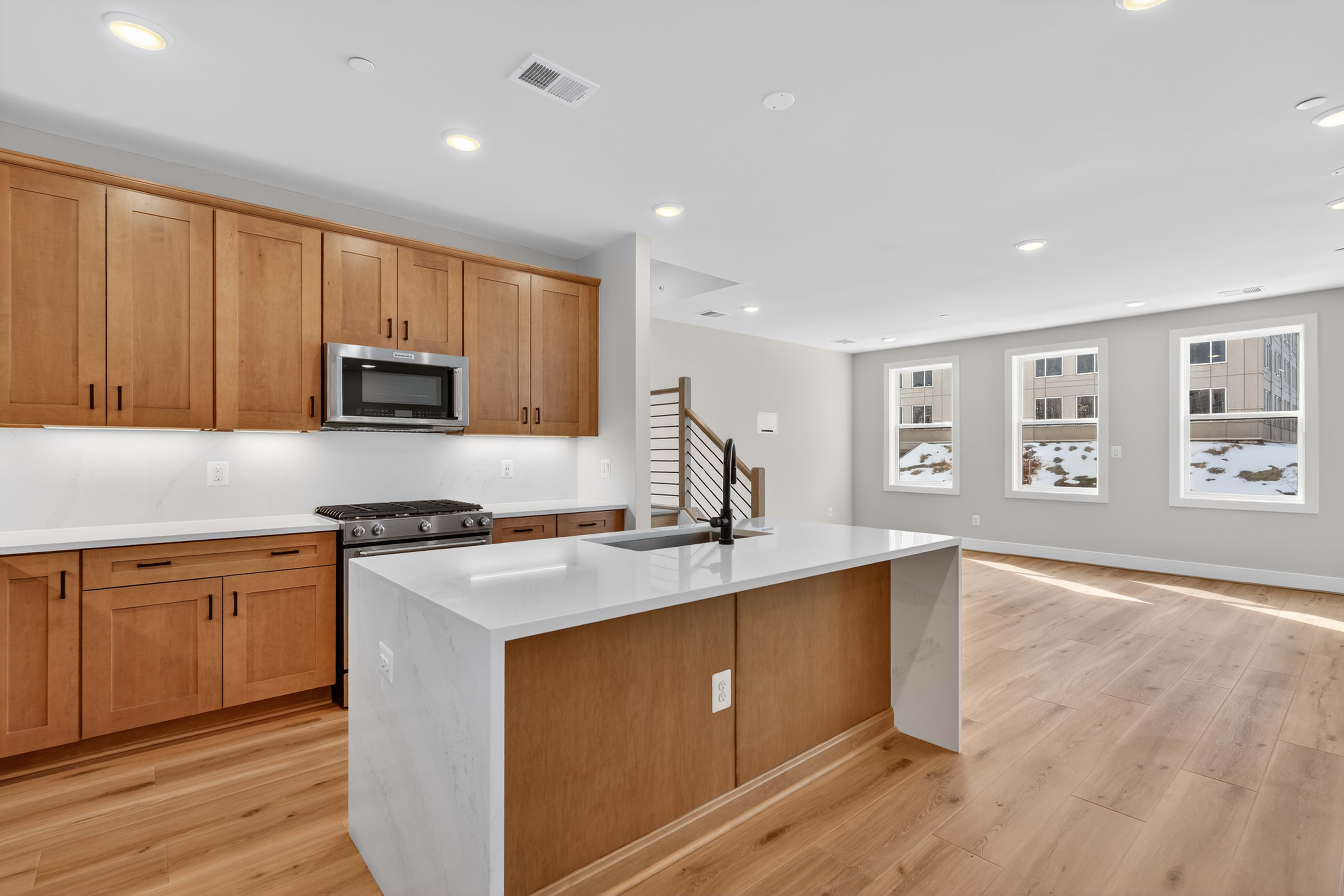 A modern, open-concept kitchen with light wood cabinets, a white countertop, and hardwood floors, with large windows providing natural light.