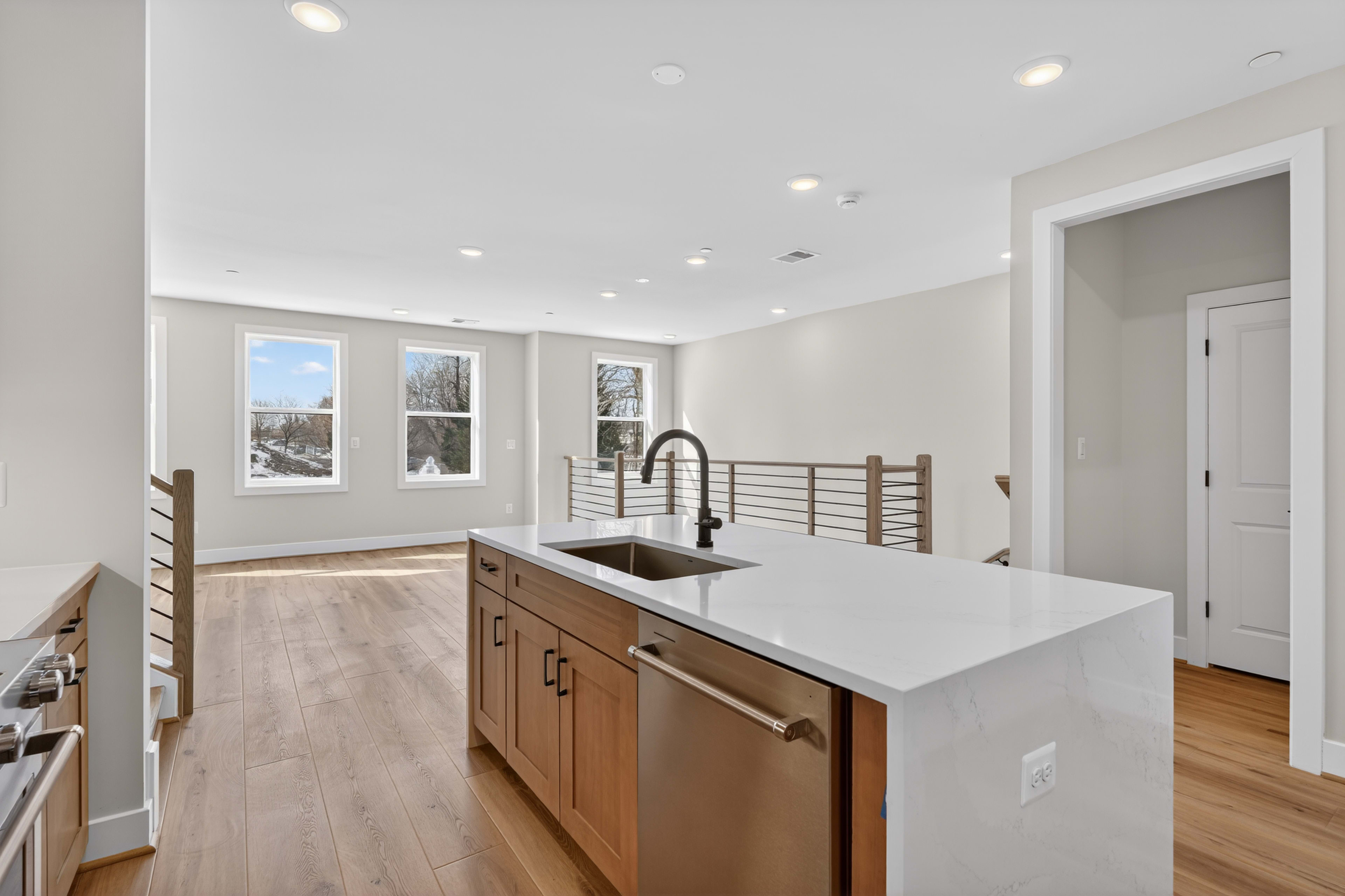 A modern, open-concept kitchen with wooden cabinets, a white countertop, and a stainless steel sink, set against a backdrop of large windows overlooking a snowy outdoor scene.