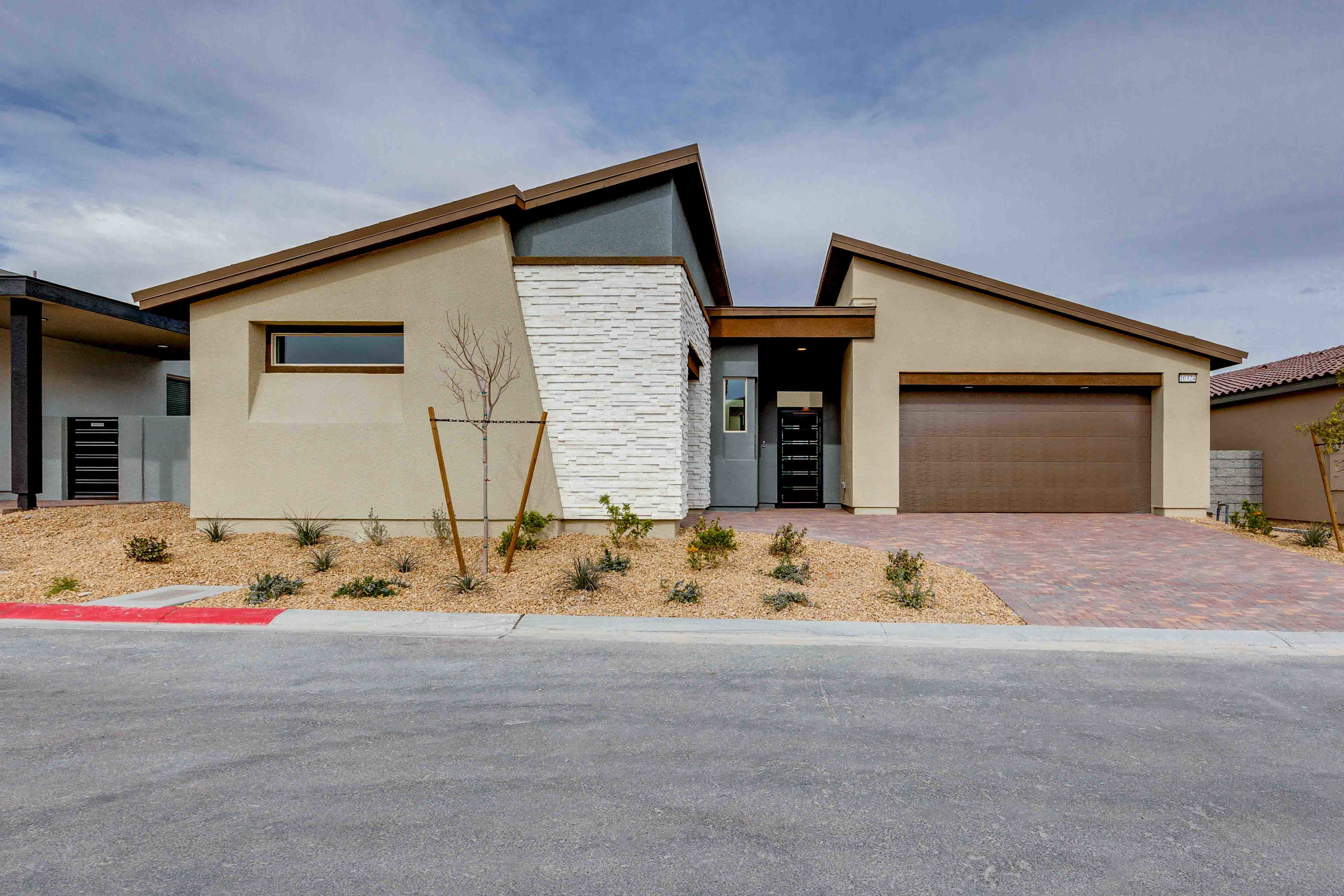 A modern, single-story house with a mix of beige and white exterior, a garage door, and a paved driveway in the foreground, set against a blue sky with scattered clouds.