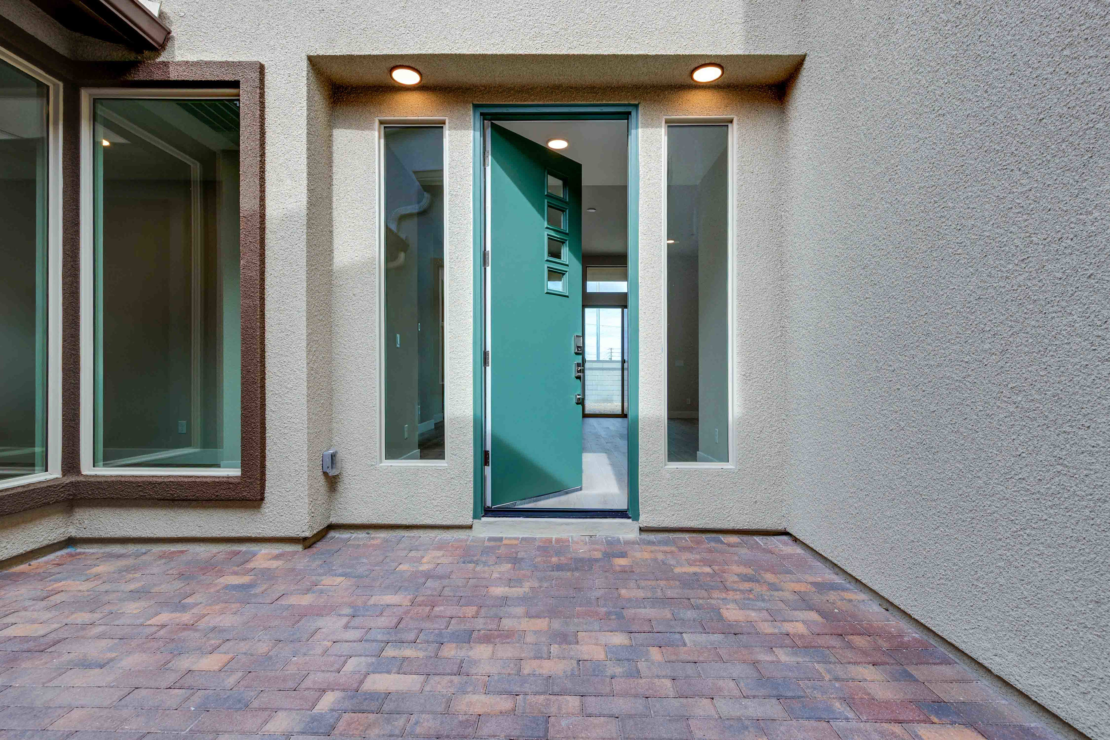 The image shows a modern entryway with a teal-colored glass door, surrounded by windows and a paved brick walkway leading up to the entrance.