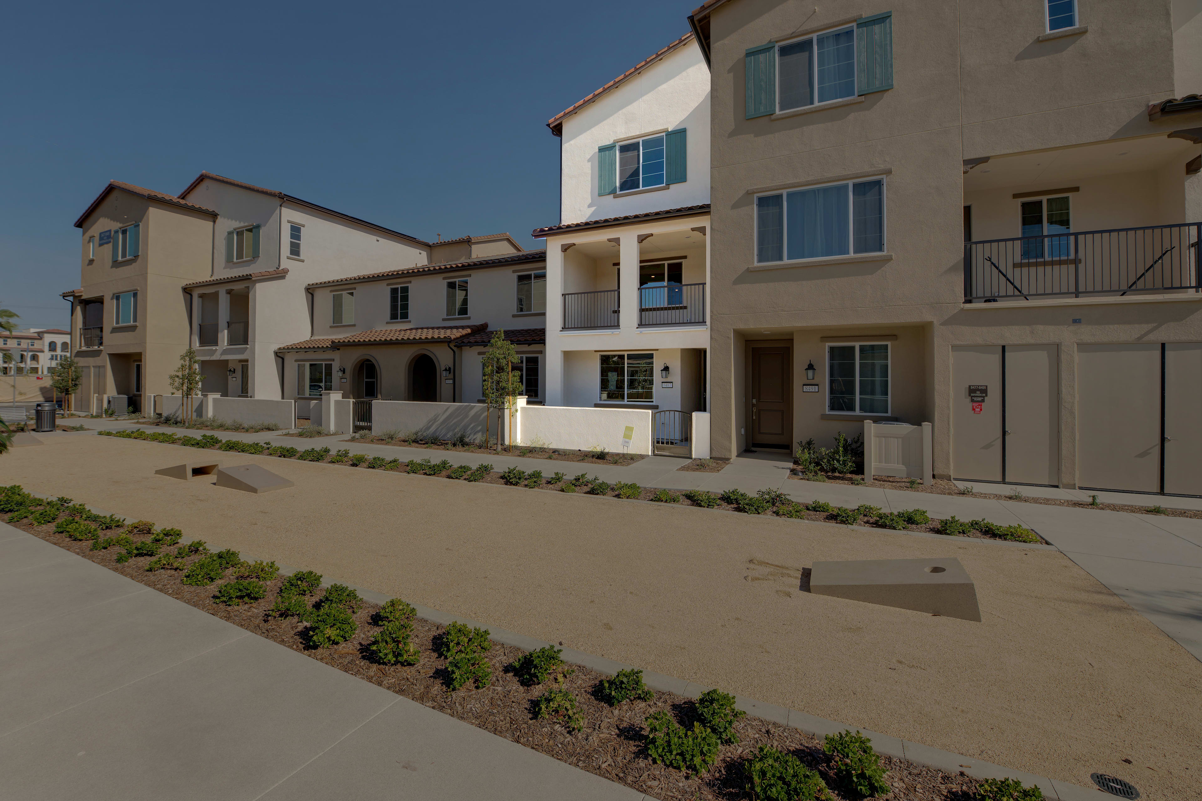 A row of multi-story residential buildings with a paved walkway and landscaped greenery in the foreground, set against a clear blue sky.