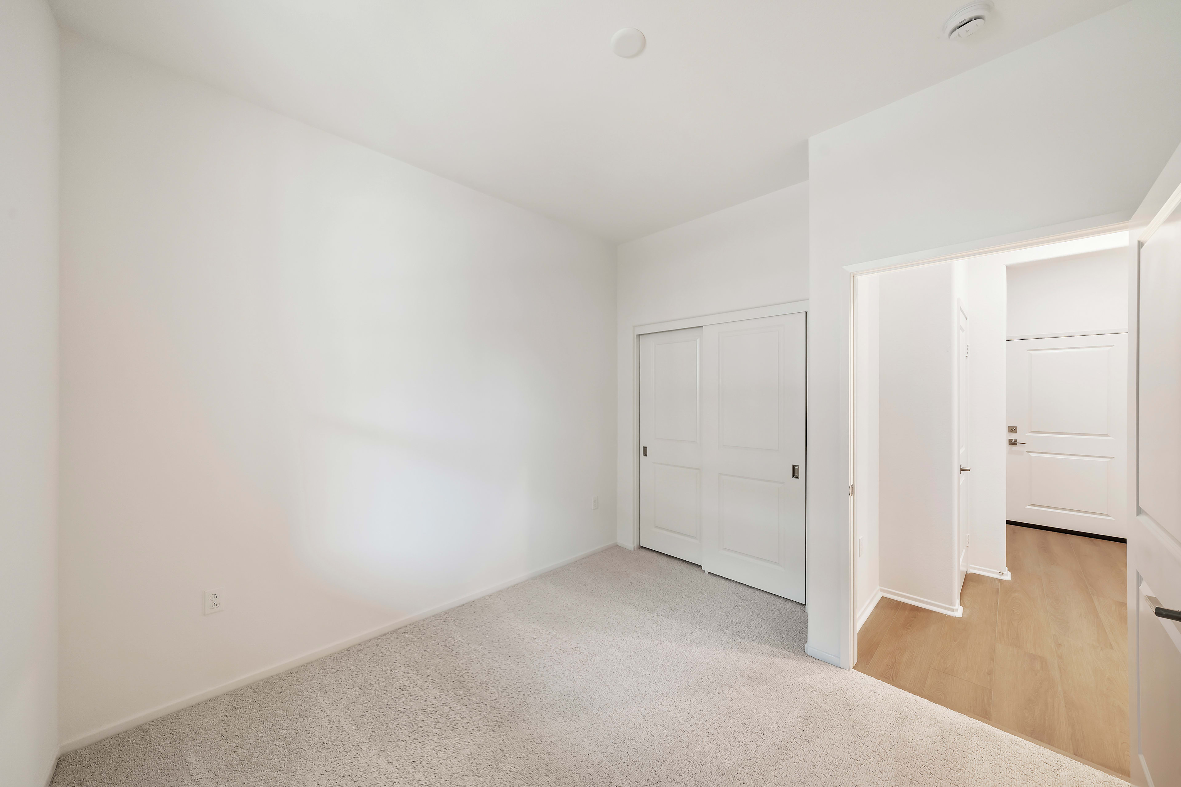 A simple, minimalist bedroom with white walls, a wooden floor, and a closed door leading to another room.