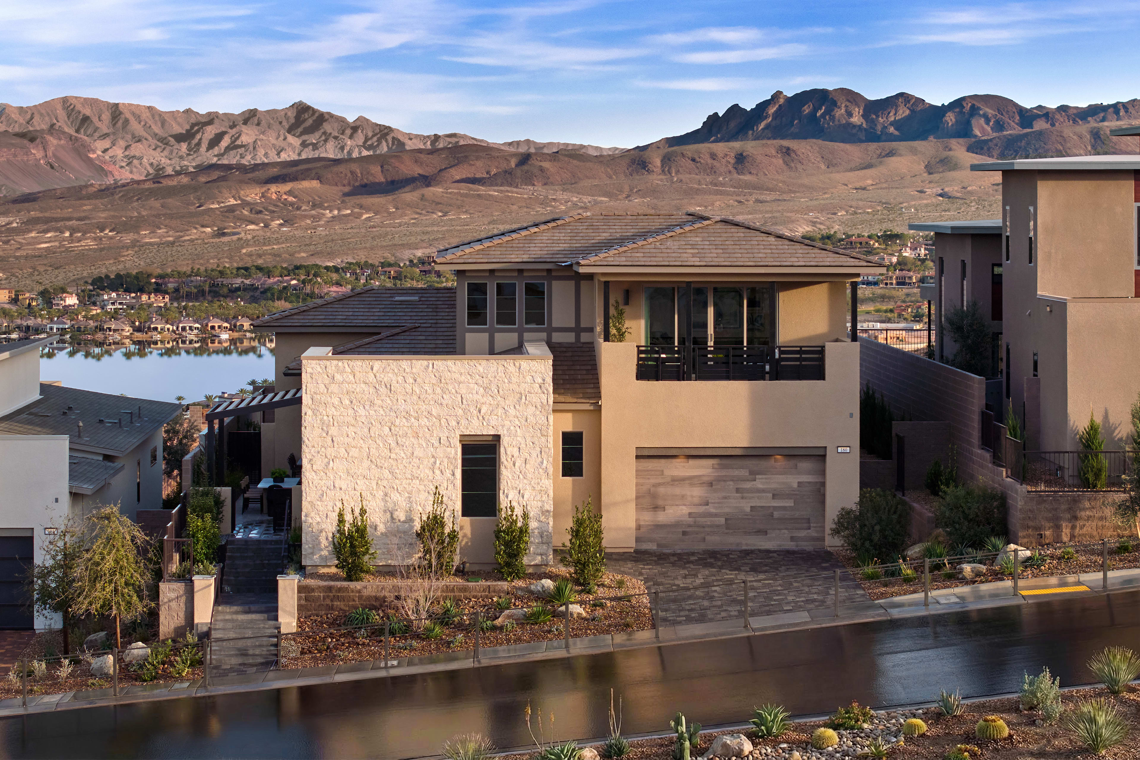 A modern, two-story house with a tile roof sits on a hillside, surrounded by a lush landscape and overlooking a scenic desert landscape with rugged mountains in the background.