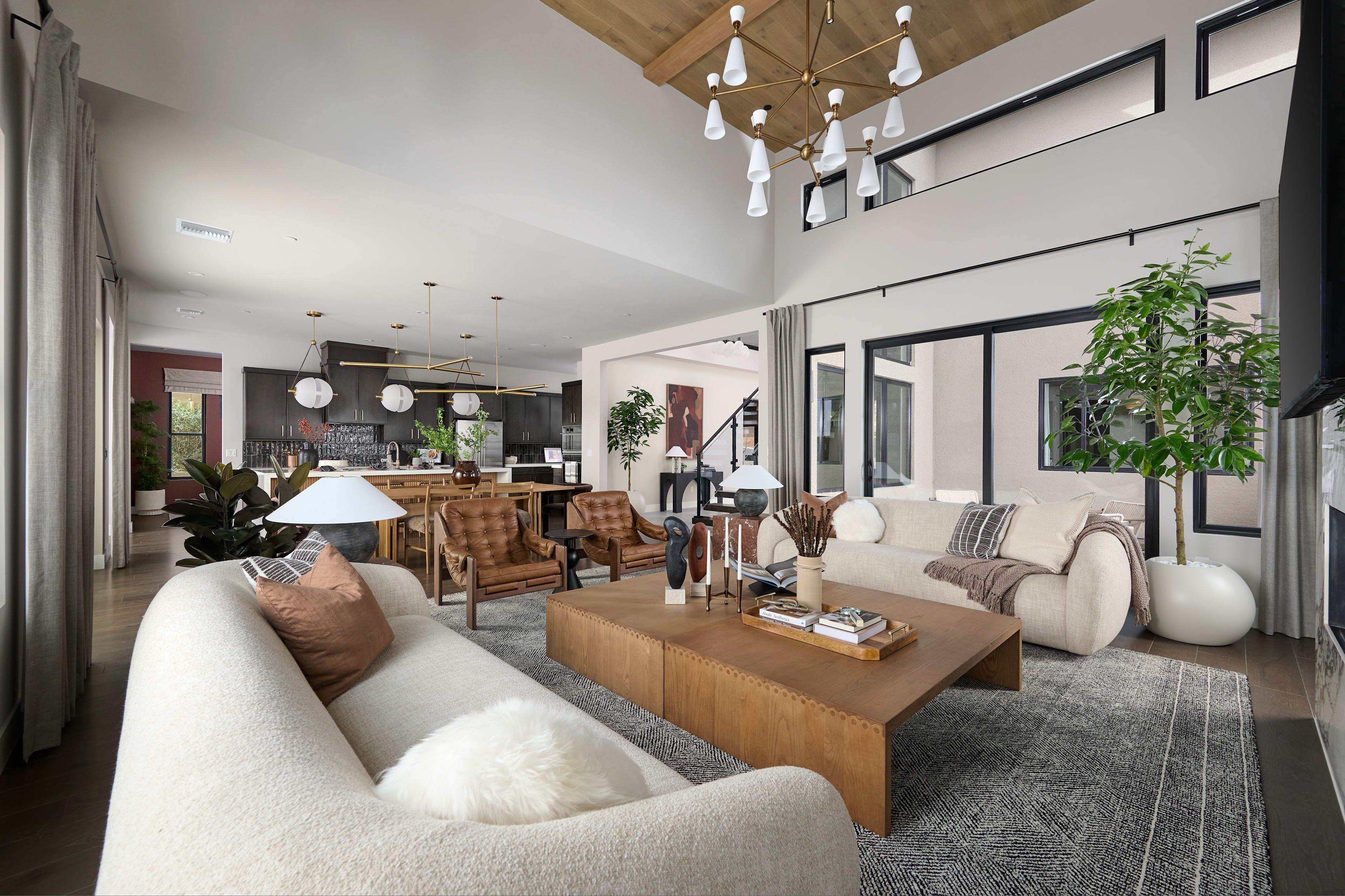 A spacious and modern living room with a cozy seating area, a wooden coffee table, and a kitchen visible in the background, all illuminated by a stylish chandelier.