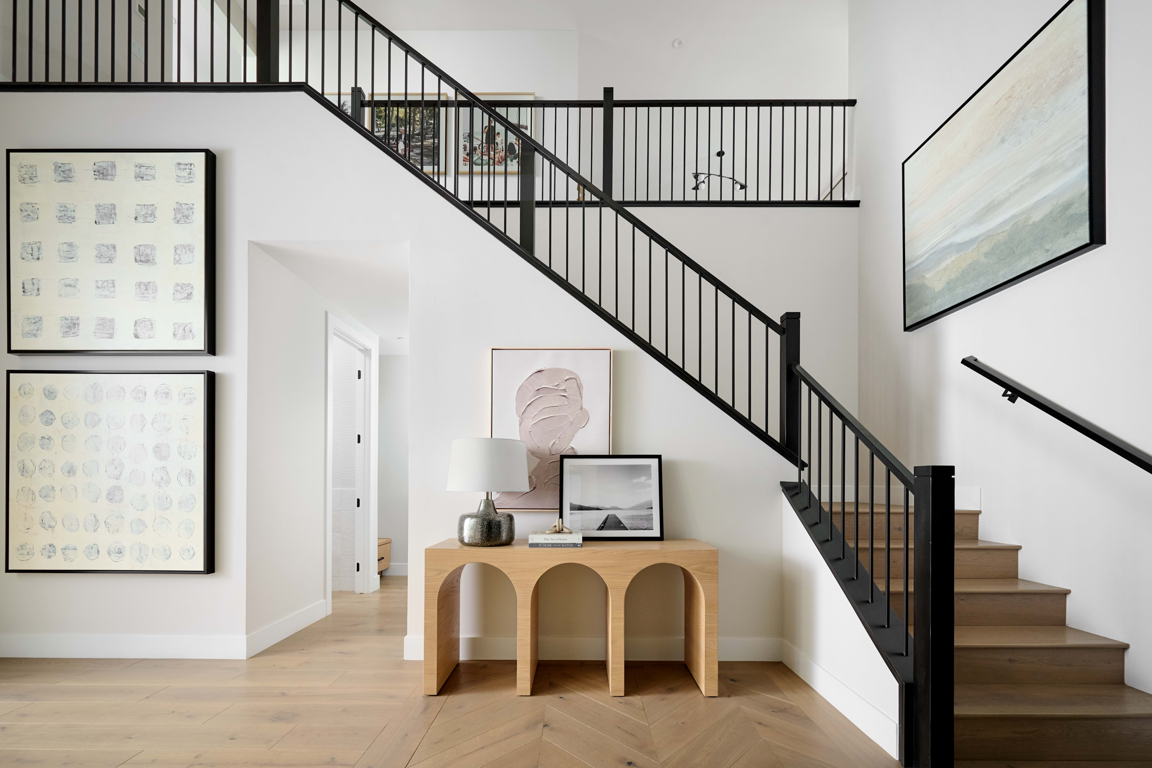 A modern, minimalist entryway with a wooden staircase, black metal railings, and various framed artwork and decor pieces on the walls.