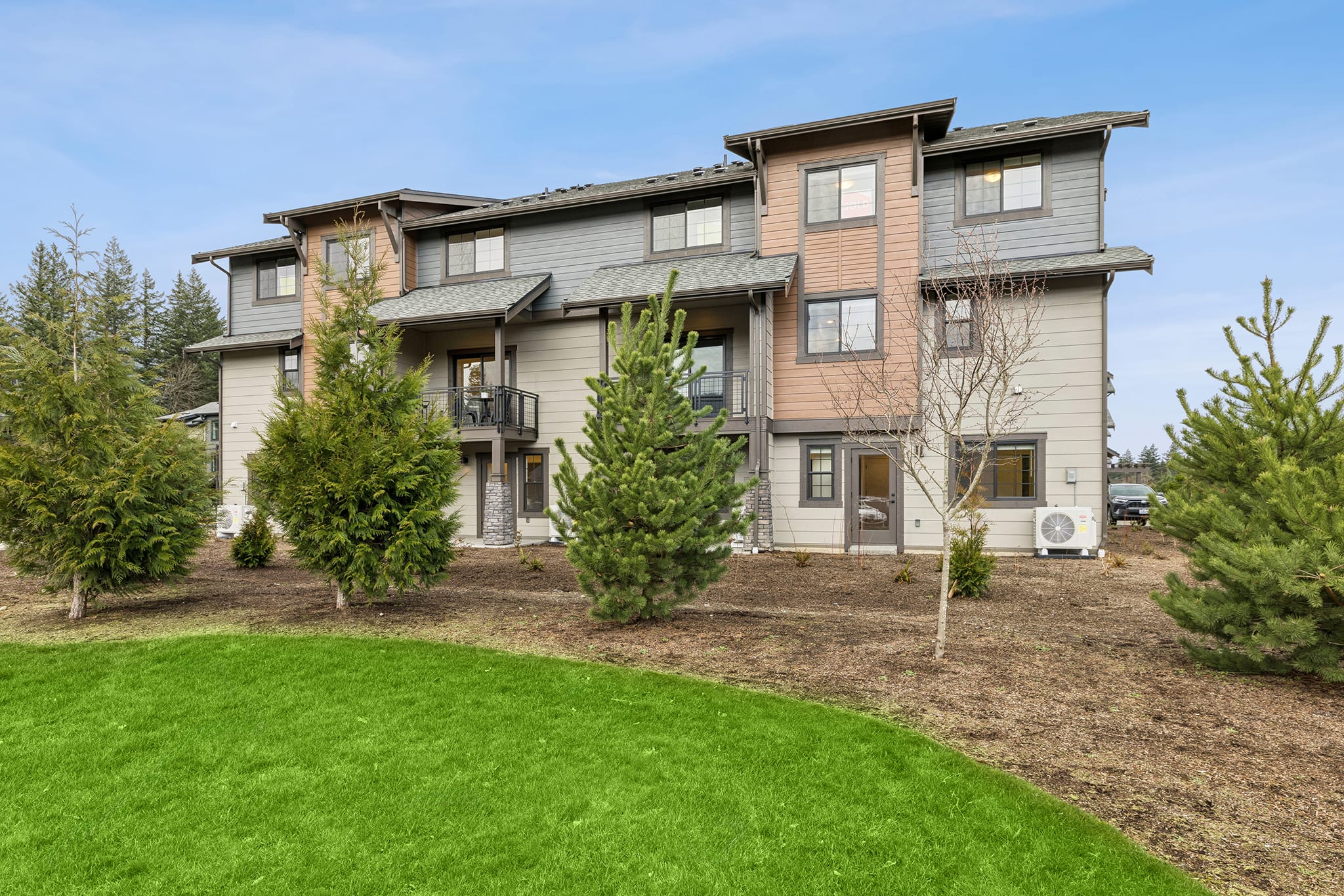 A multi-story residential building with a well-manicured lawn and landscaping in the foreground, surrounded by trees and a clear blue sky in the background.