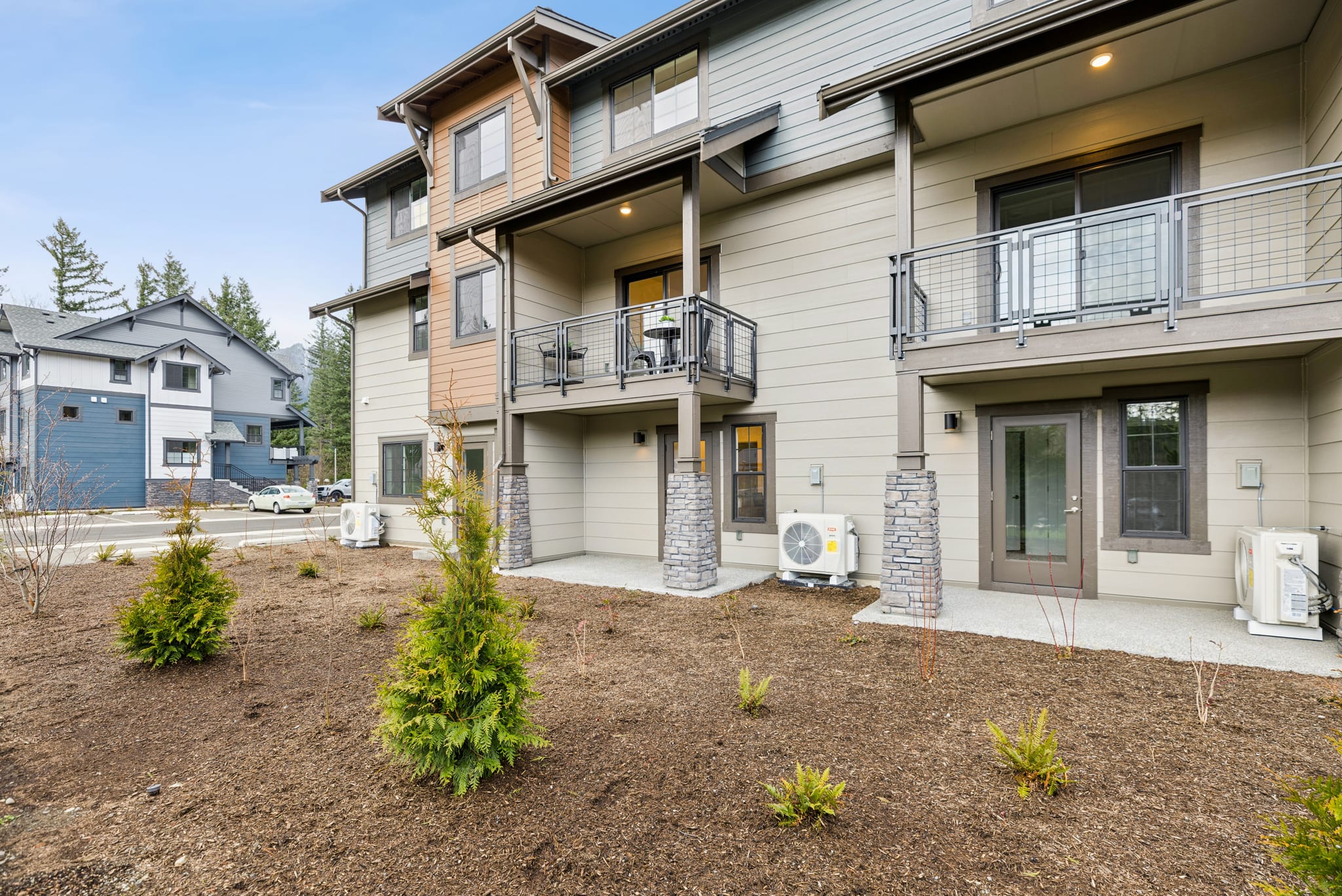 A multi-story residential building with a landscaped front yard, featuring a mix of evergreen and deciduous plants, and a paved driveway leading to the building's entrance.