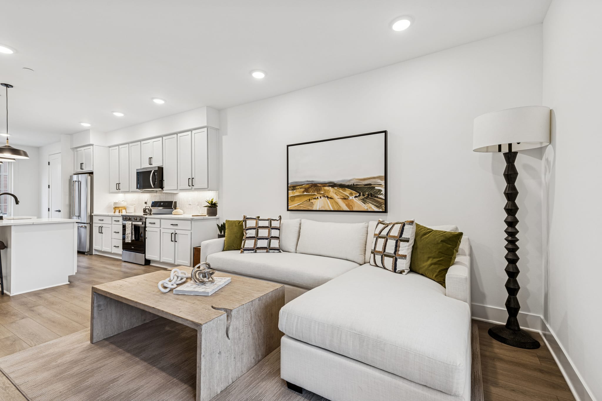A modern and cozy living room with a white sofa, a wooden coffee table, and a large framed artwork on the wall, complemented by a kitchen area with white cabinets and stainless steel appliances.