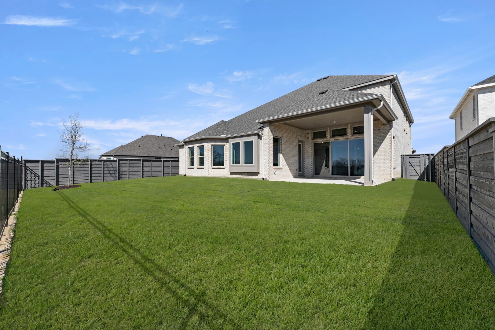 A well-manicured lawn surrounds a modern, two-story house with a covered porch and large windows, set against a clear blue sky.