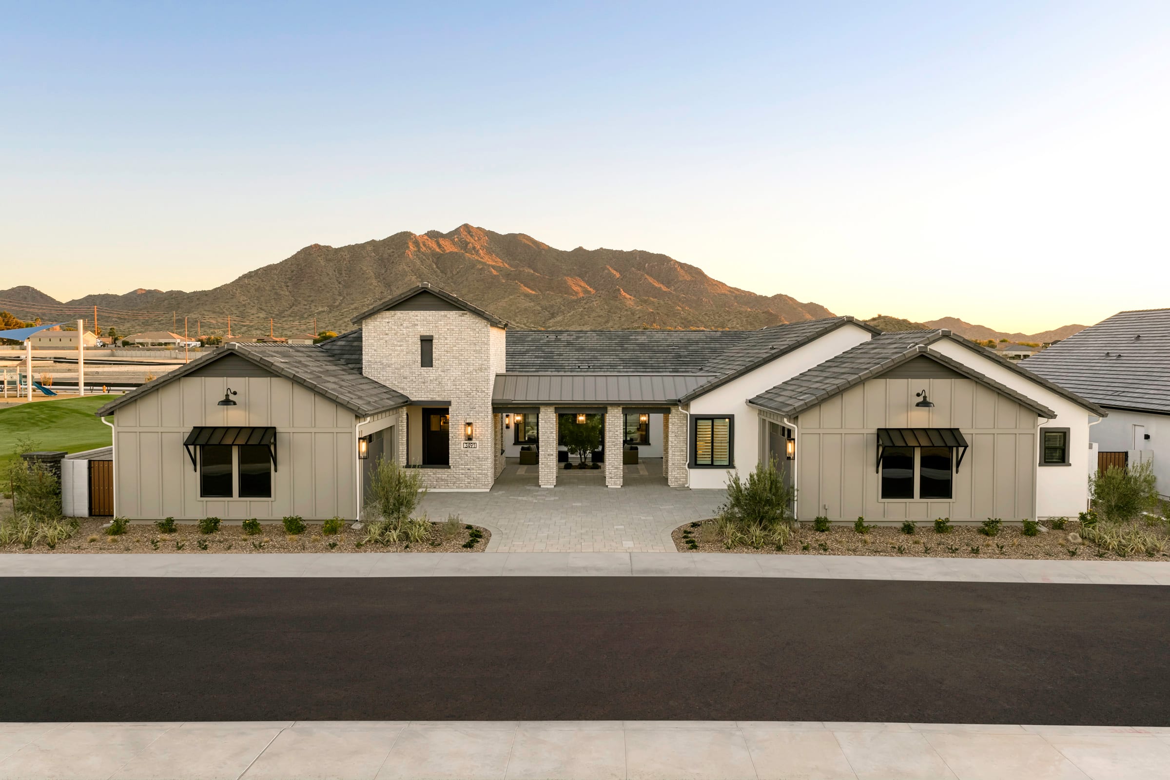 A modern farmhouse-style building with a central tower and two wings sits in the foreground, surrounded by a grassy field. In the background, rugged mountains rise against a clear blue sky.