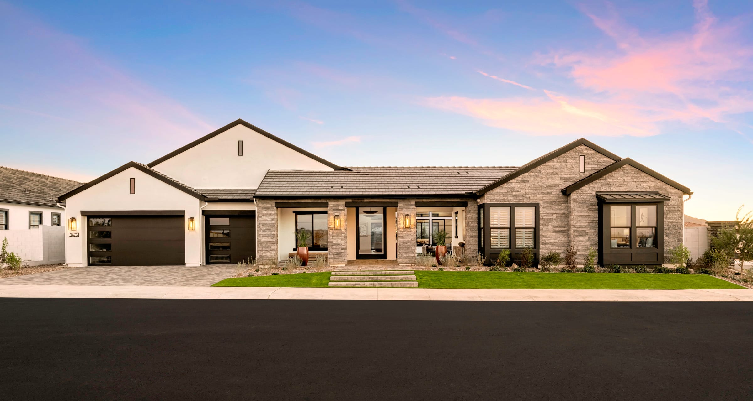 A modern, two-story house with a stone exterior, a tiled roof, and a well-manicured lawn in the foreground, set against a vibrant sky with pastel-colored clouds at sunset.