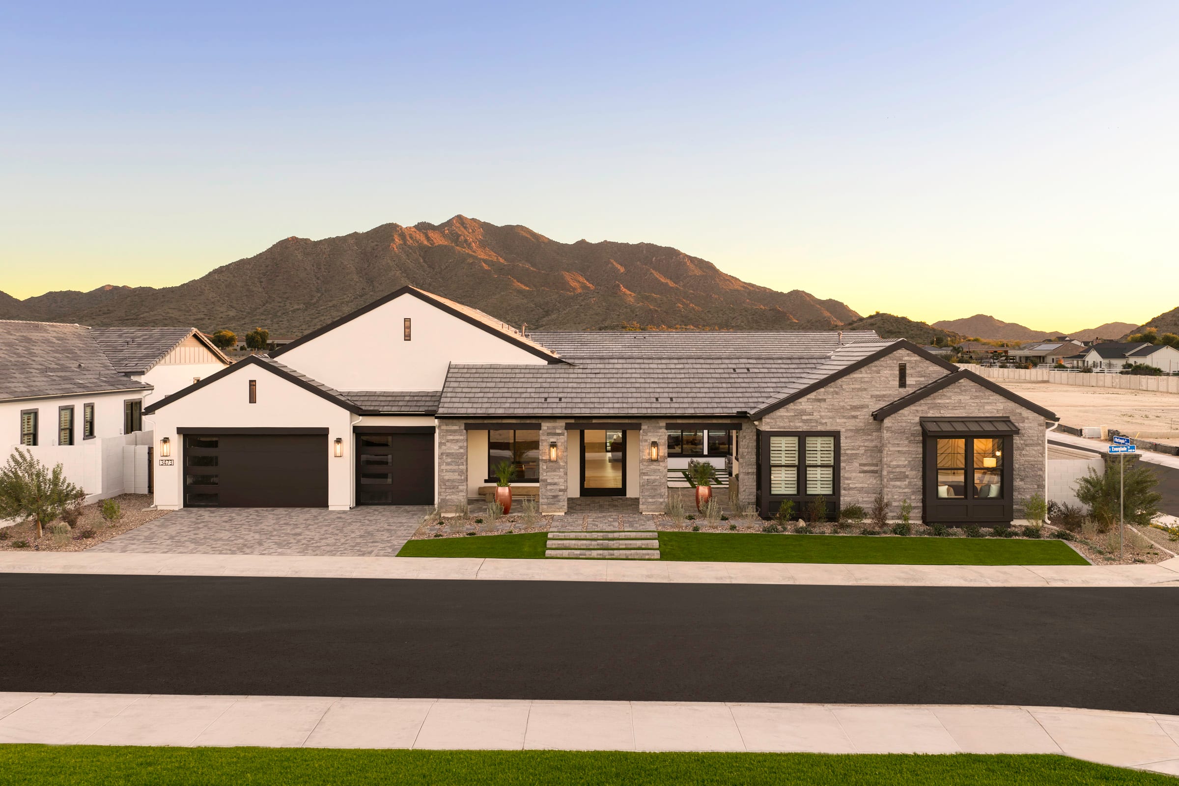 A modern, single-story house with a stone exterior and a tiled roof sits in the foreground, surrounded by a well-manicured lawn. In the background, rugged mountains rise against a clear, blue sky, creating a picturesque desert landscape.