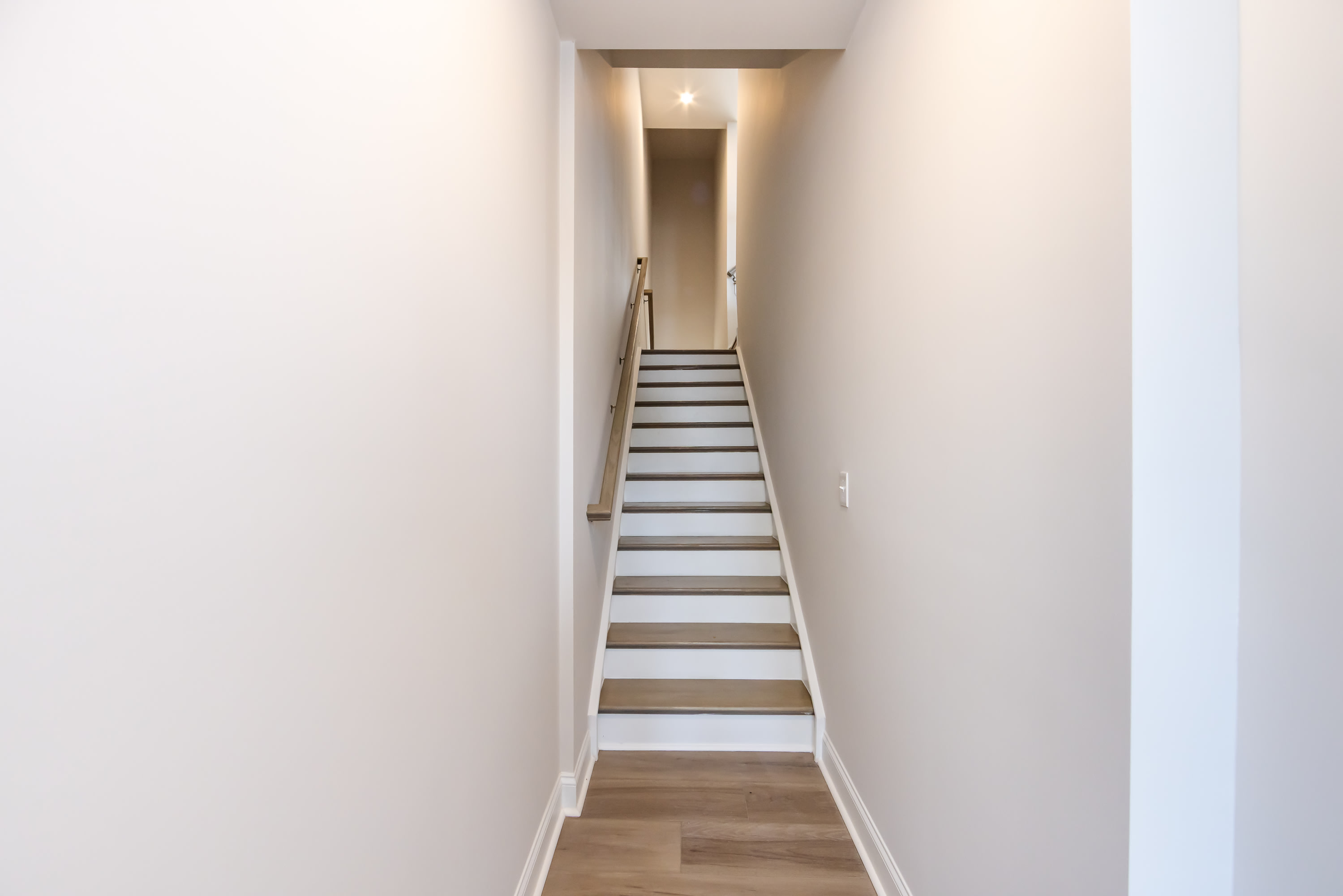 A narrow staircase with wooden steps leads up to a brightly lit hallway, with white walls and a wooden floor visible in the background.