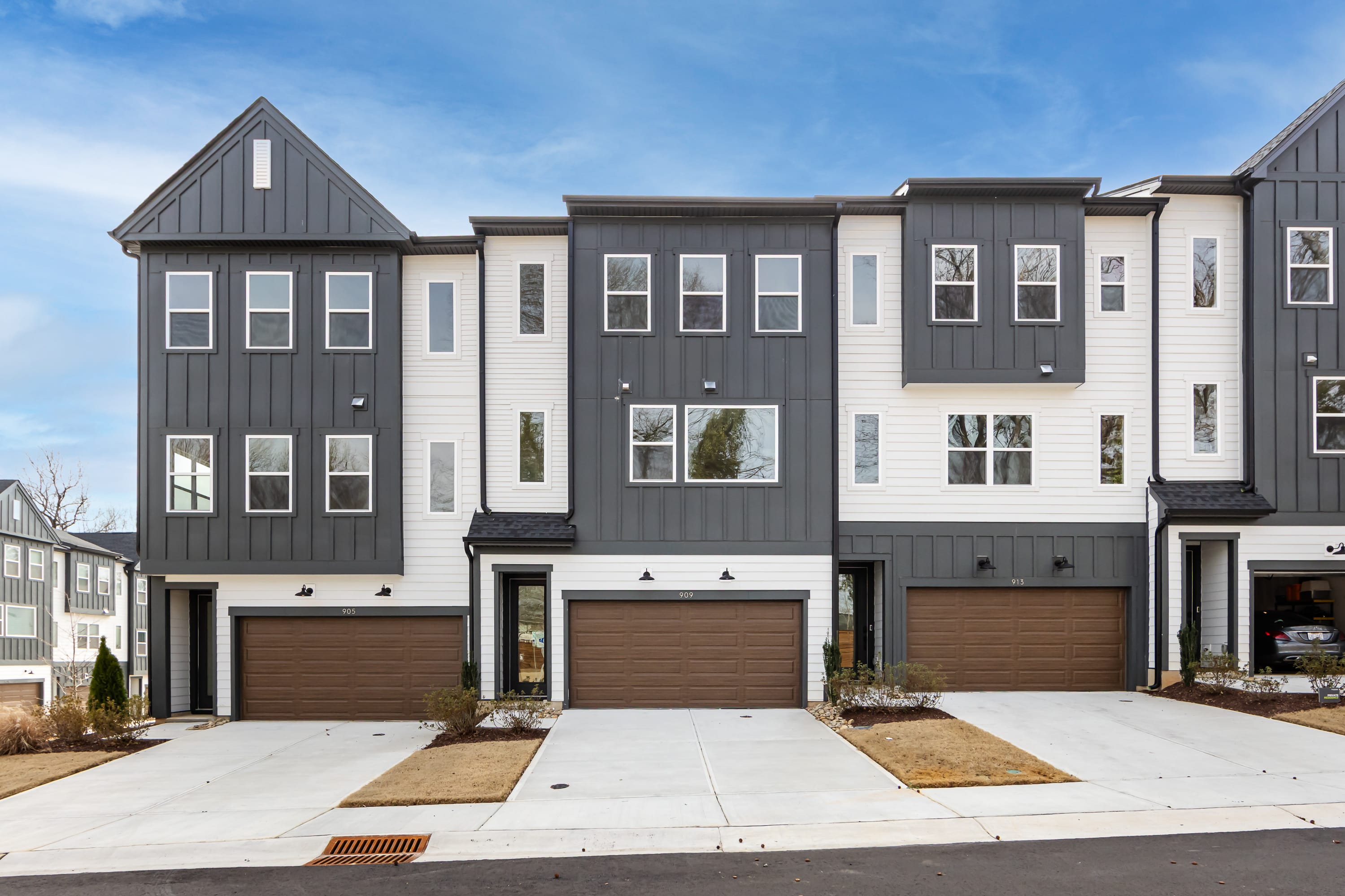 A row of modern, multi-story townhouses with gray siding, white trim, and garage doors in the foreground, set against a clear blue sky in the background.