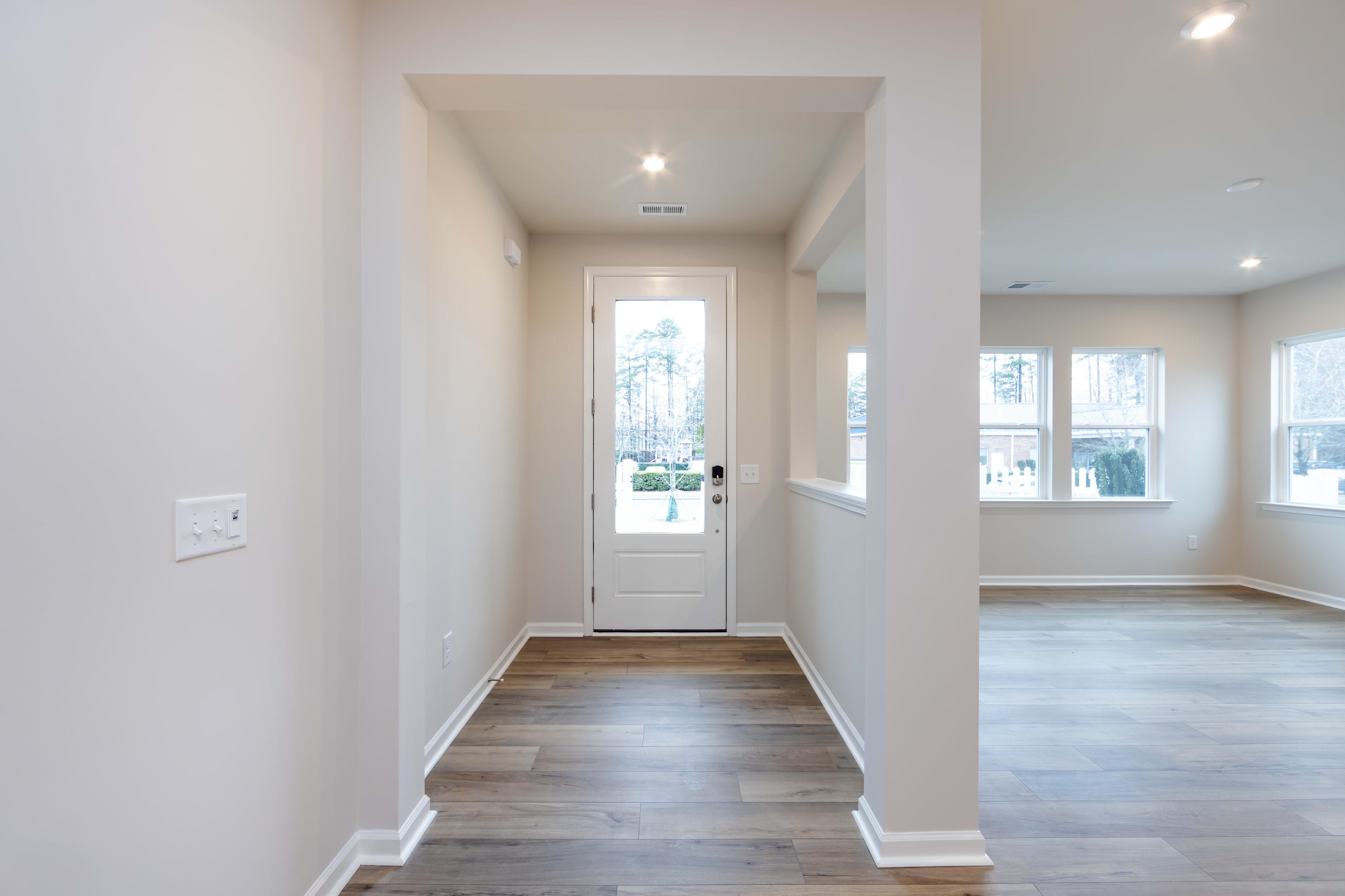 A bright and spacious entryway with a white door leading to the outdoors, hardwood floors, and recessed lighting illuminating the clean, minimalist interior.