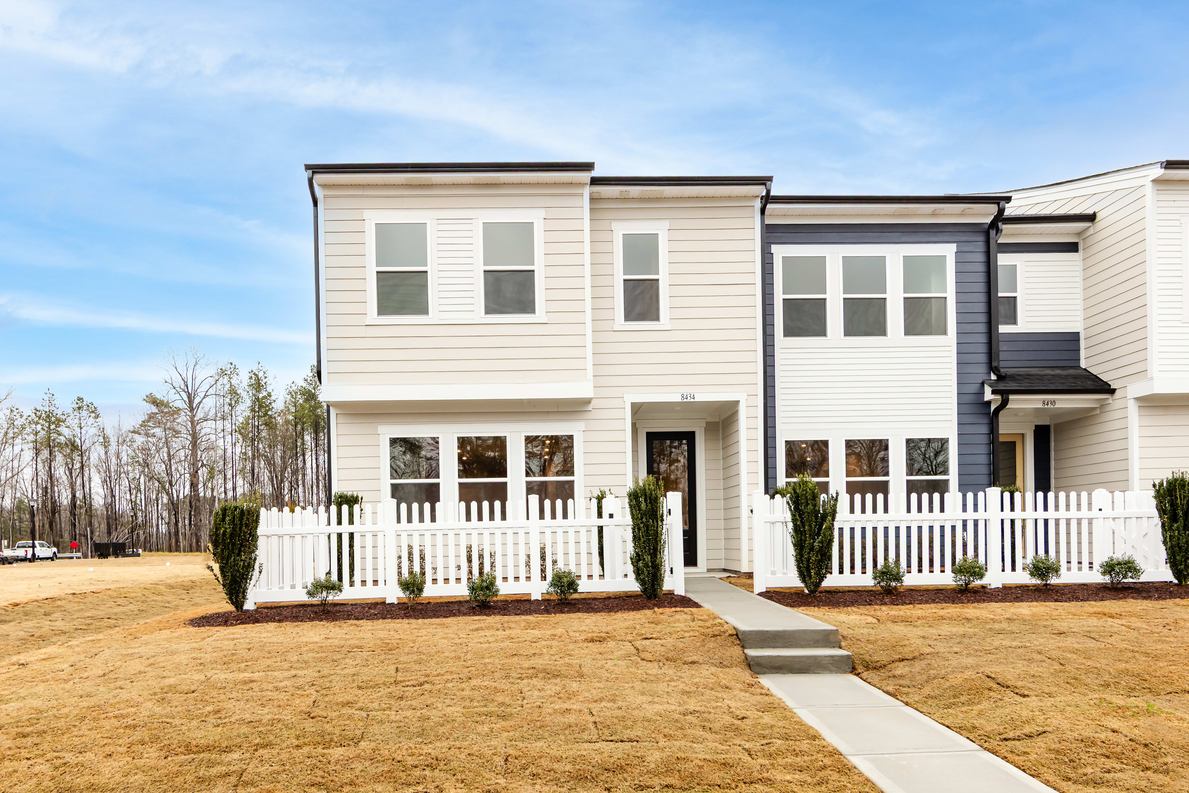 A modern two-story townhouse with a white exterior, a porch, and a well-manicured lawn surrounded by trees in the background.