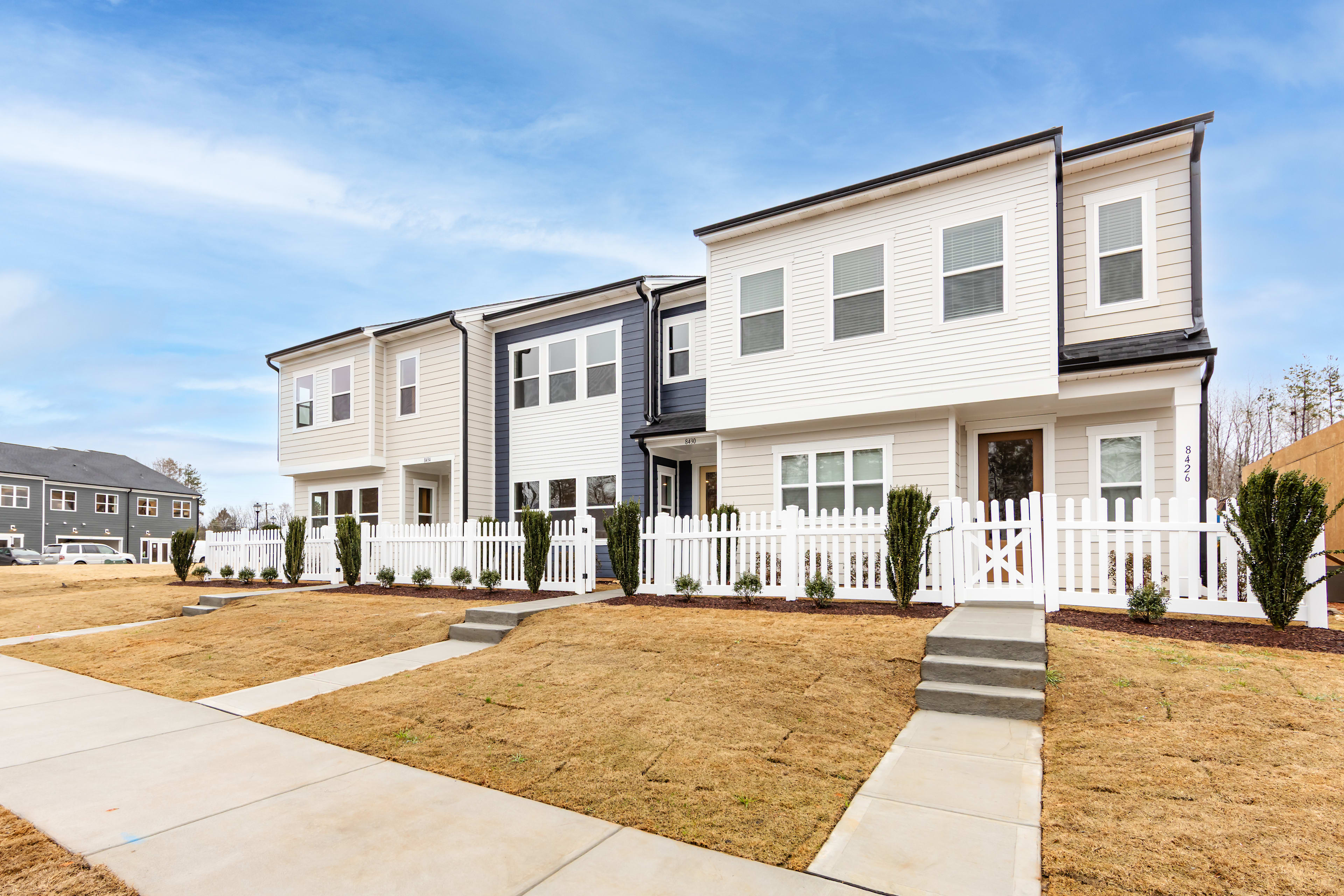A row of modern, two-story townhouses with white siding, black trim, and fenced yards sits in a residential neighborhood with a clear blue sky overhead.