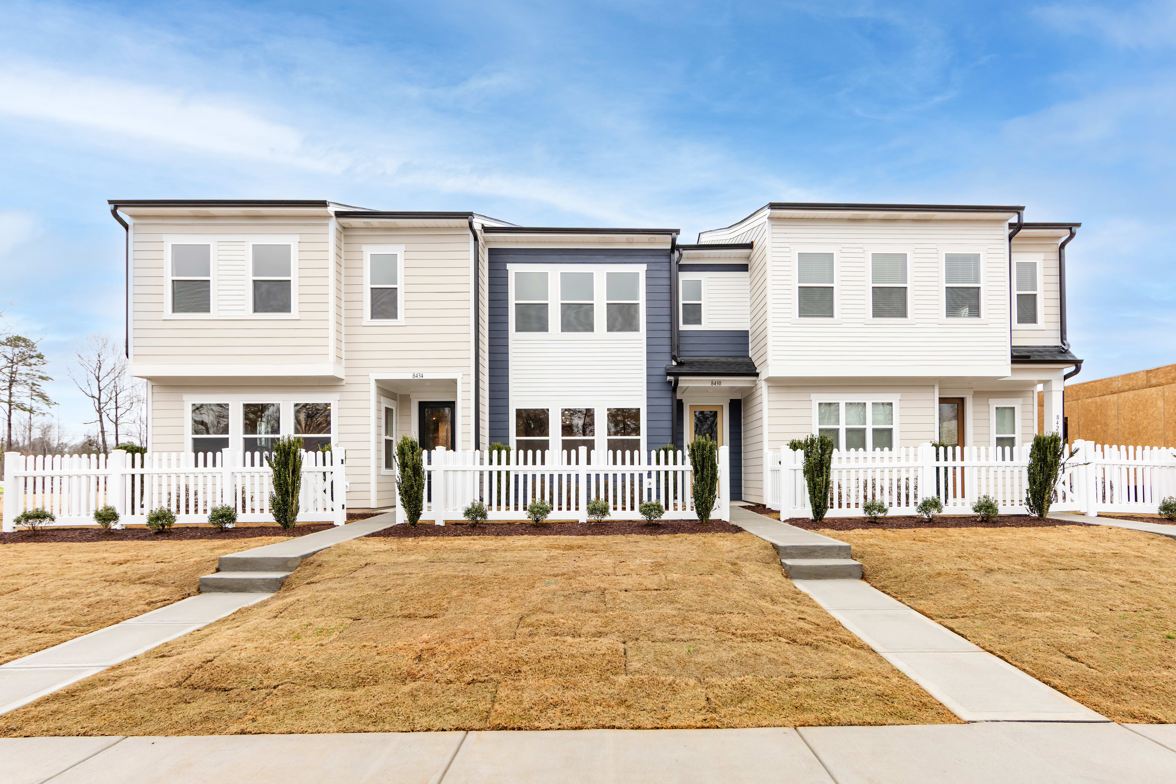 A modern, two-story townhouse community with a well-manicured lawn and a white picket fence in the foreground, set against a clear blue sky.
