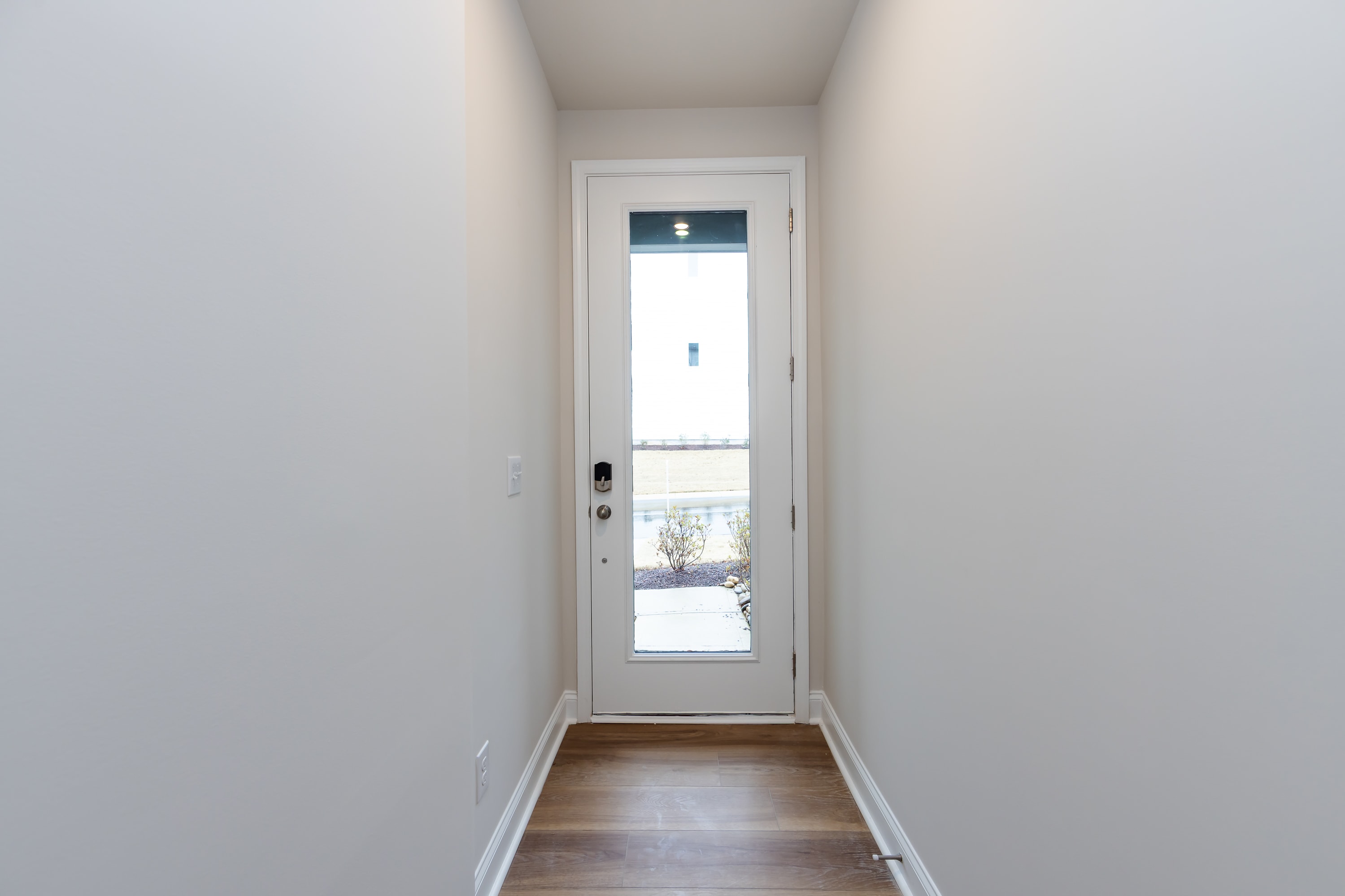 A narrow hallway with a white door leading to the outside, featuring a wooden floor and plain white walls.