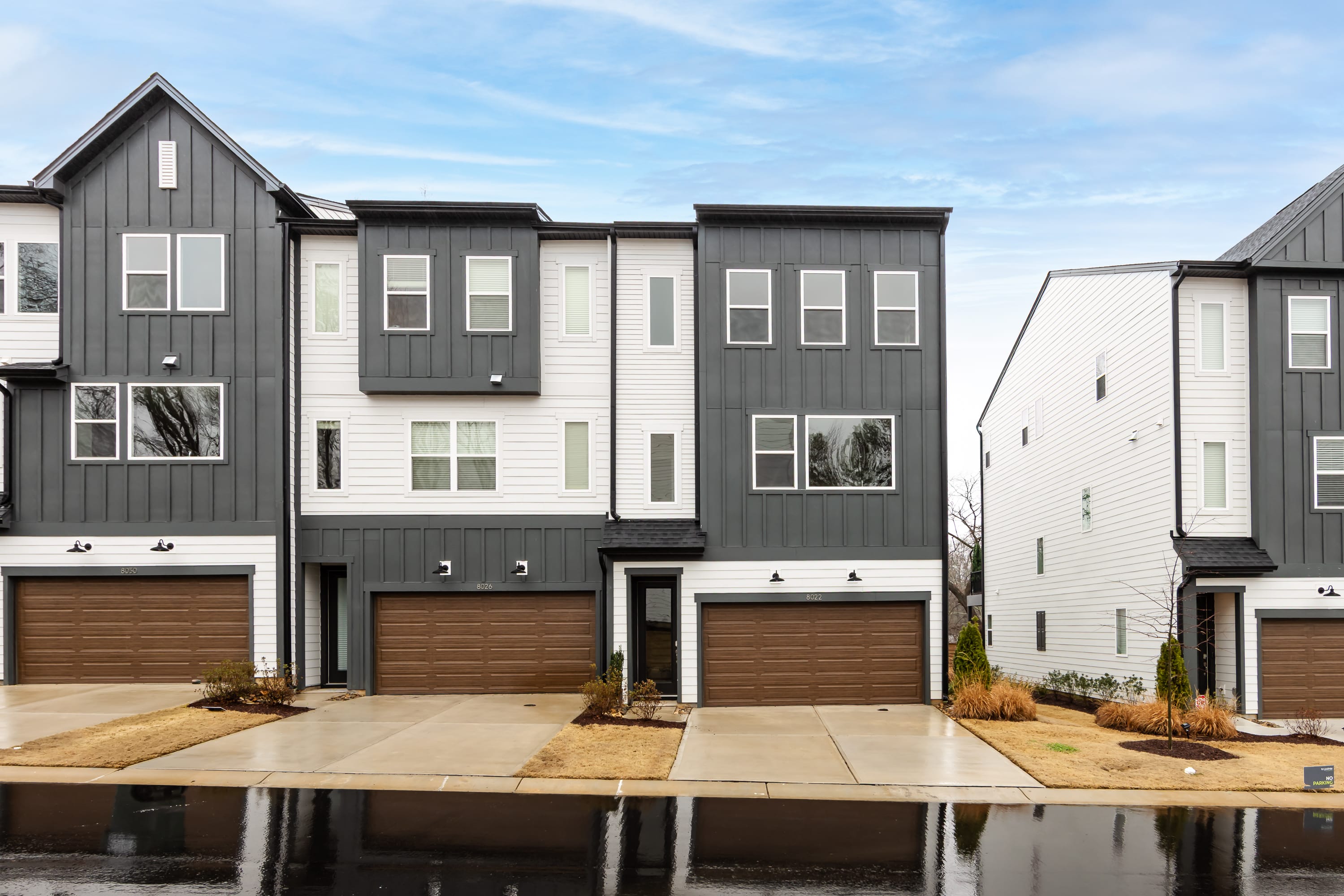 A row of modern, multi-story townhouses with gray siding, large windows, and attached garages, set against a blue sky with wispy clouds.