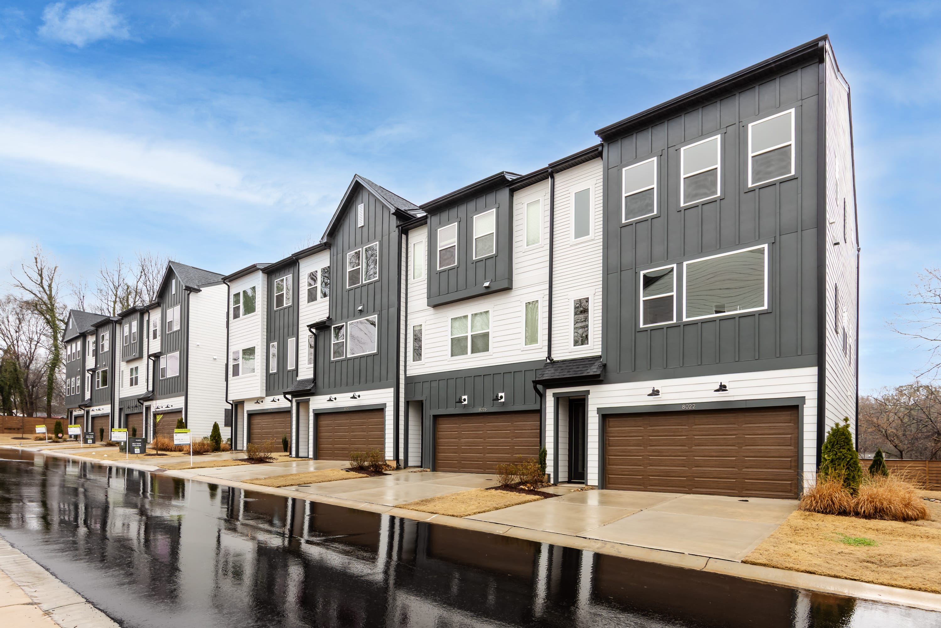 A row of modern, multi-story townhouses with gray siding and garages, reflected in a puddle in the foreground against a blue sky.